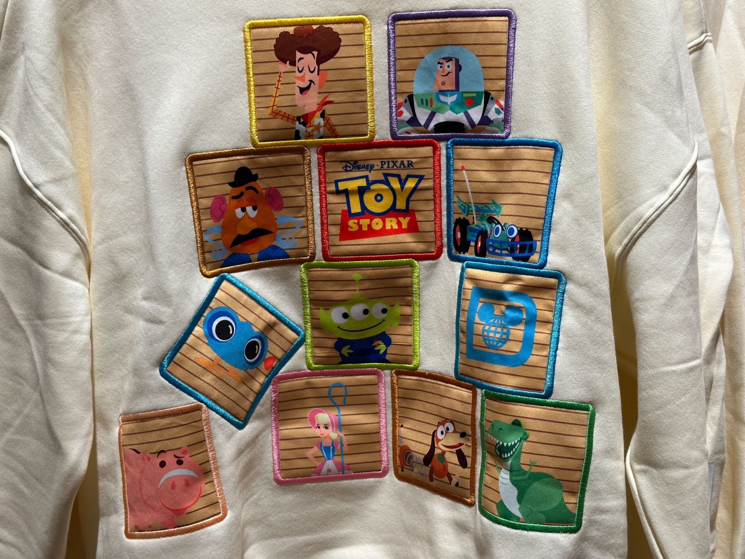 A sweatshirt displays colorful Toy Story character patches circling the logo, evoking playful vibes like in Disney park merchandise.