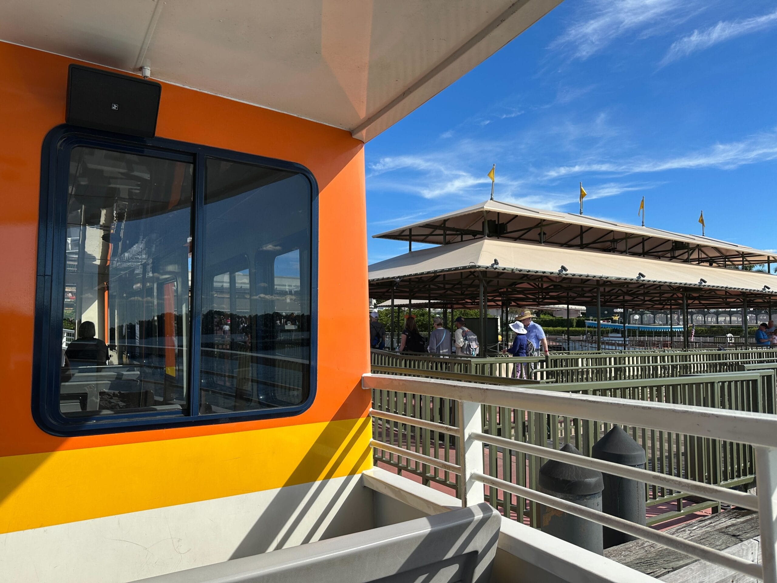A ferry with orange and yellow stripes, reminiscent of Disney transportation, is docked as guests wait under clear blue skies.