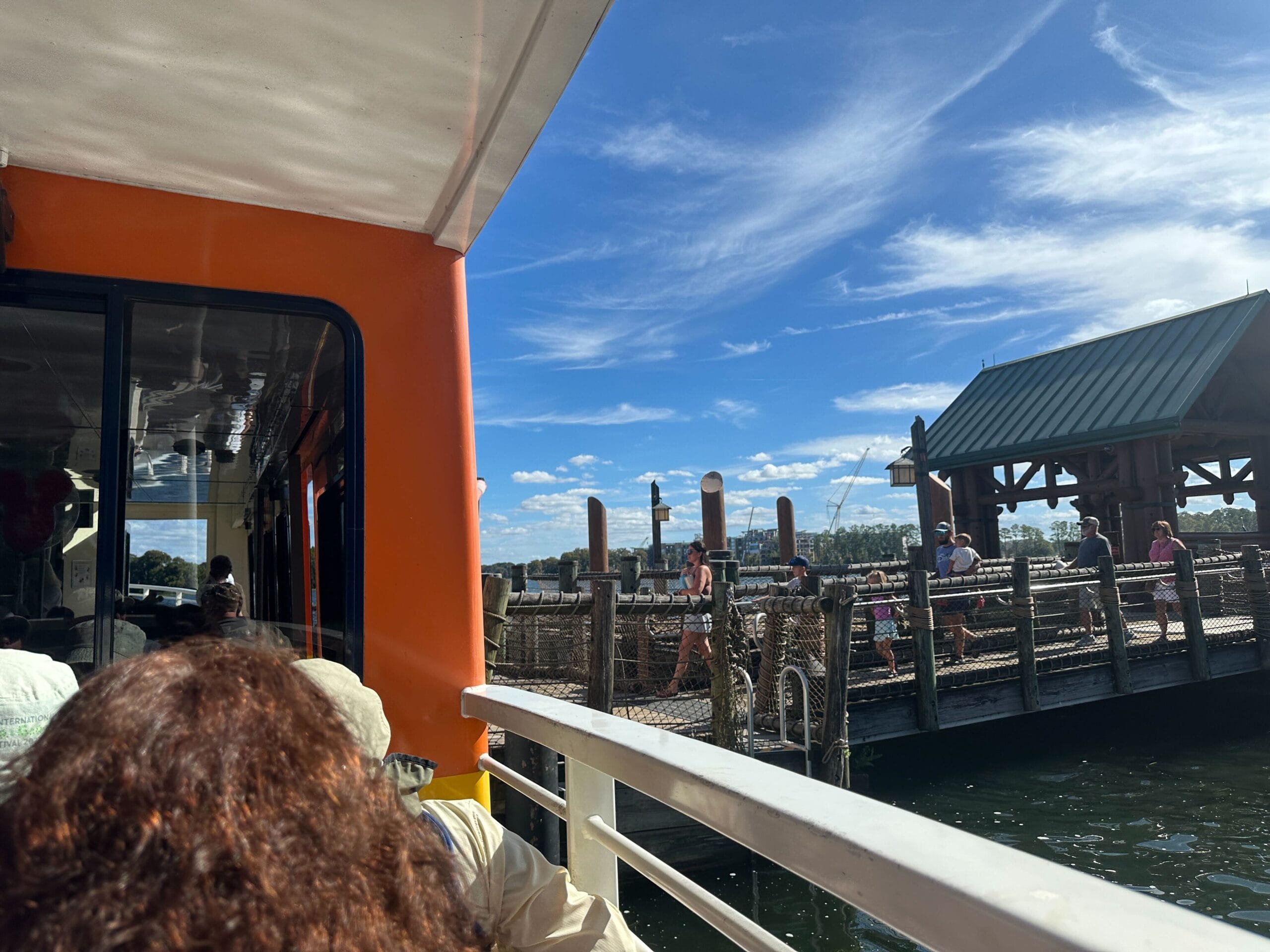 Guests ride a boat approaching a themed dock at a Disney park, beneath a blue sky with scattered clouds.