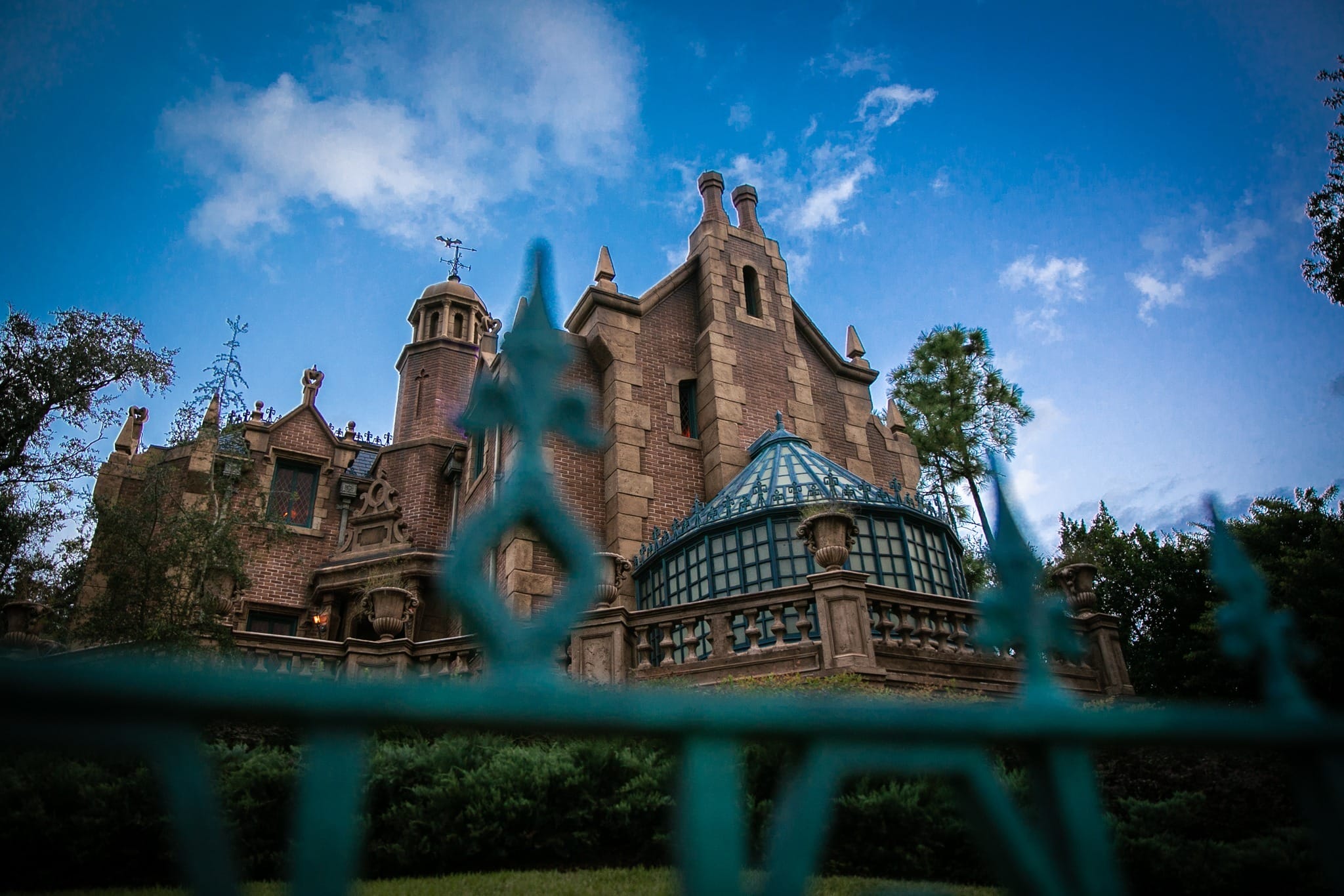 The Haunted Mansion with eerie gothic architecture stands behind a blue iron fence, evoking the classic Disney park attraction.