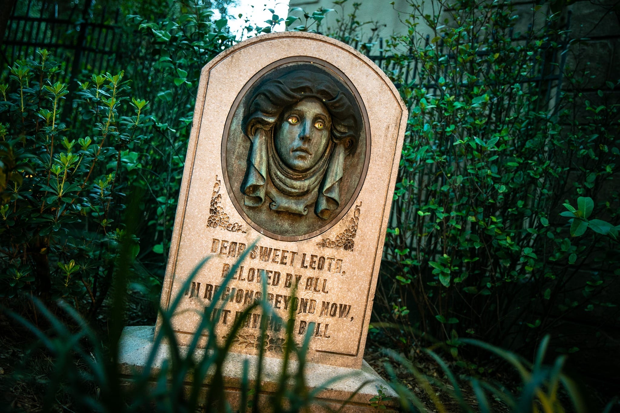 A tilted gravestone with a glowing-eyed woman’s face, echoing Disney’s Haunted Mansion, rests in a lush garden setting.