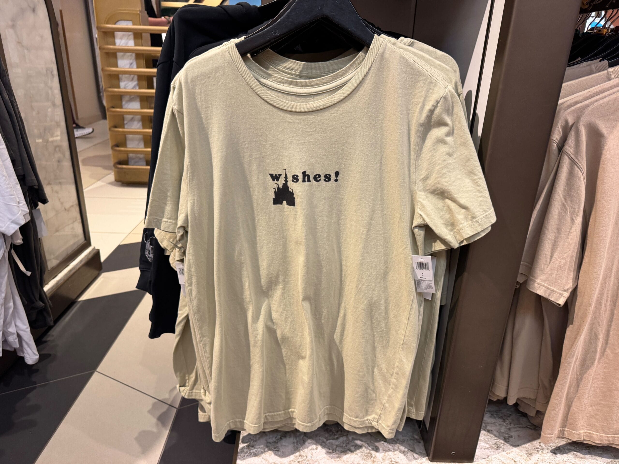 Beige T-shirt on a hanger with "Wishes!" in black text, displayed in a Disney park shop, reminiscent of fireworks shows at Magic Kingdom.