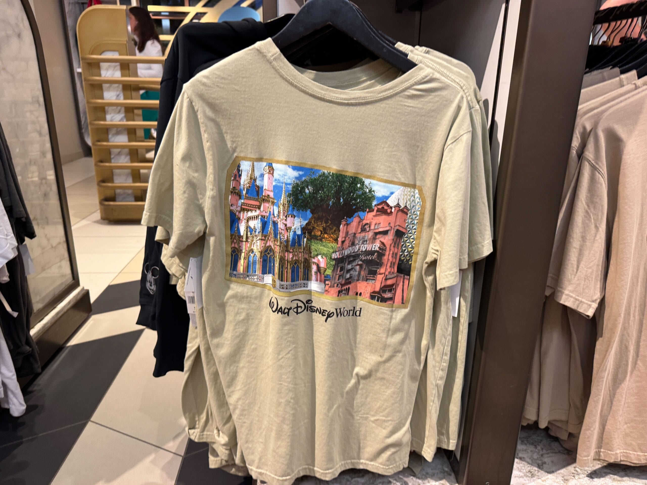 Beige T-shirt with a Walt Disney World graphic featuring Cinderella Castle, modeled after Magic Kingdom’s, plus park icons, on display.