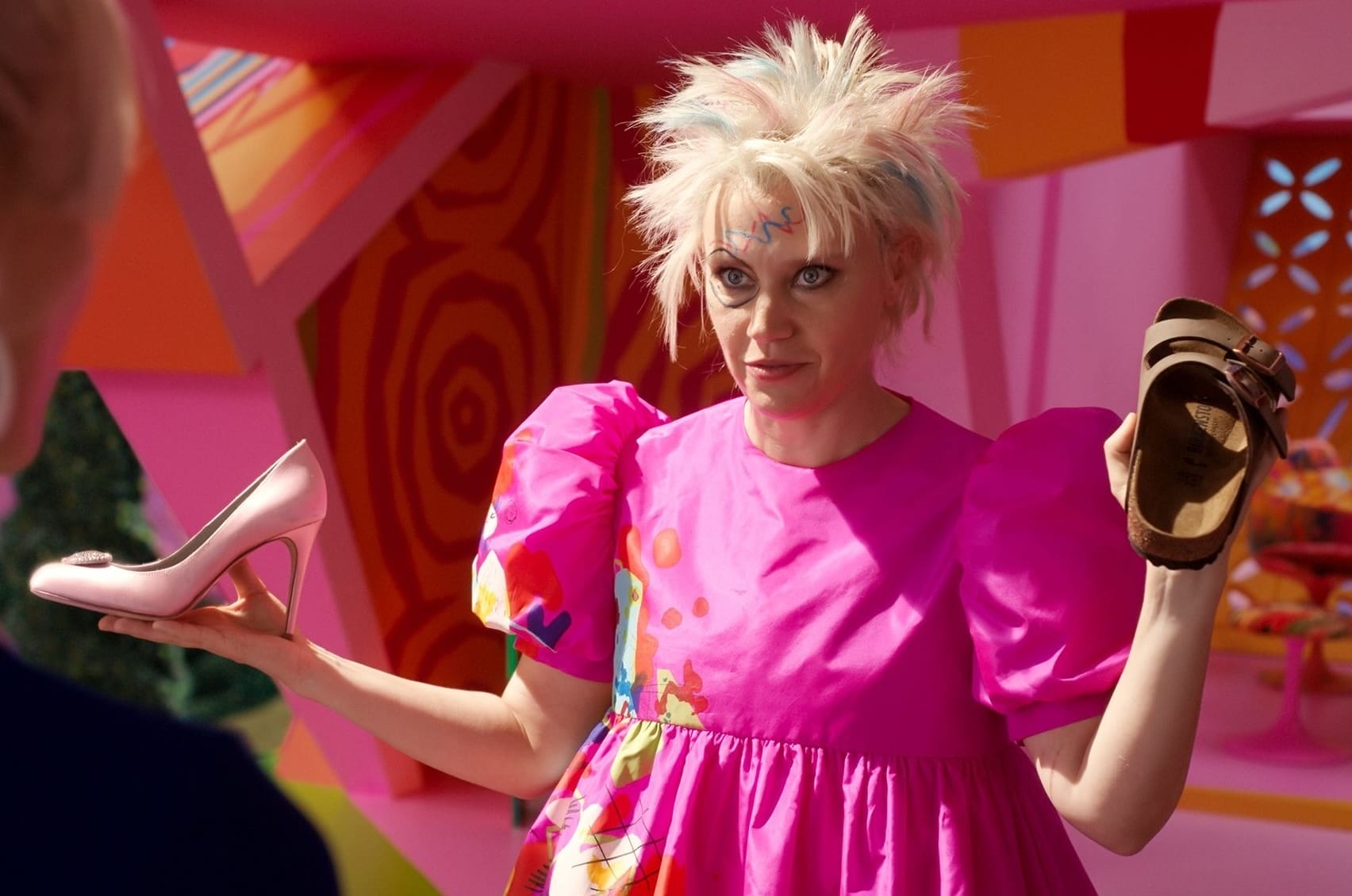 Kate McKinnon as Weird Barbie in the Barbie movie in her paint-splattered pink dress holding a pink high-heeled shoe and a flat brown sandal.