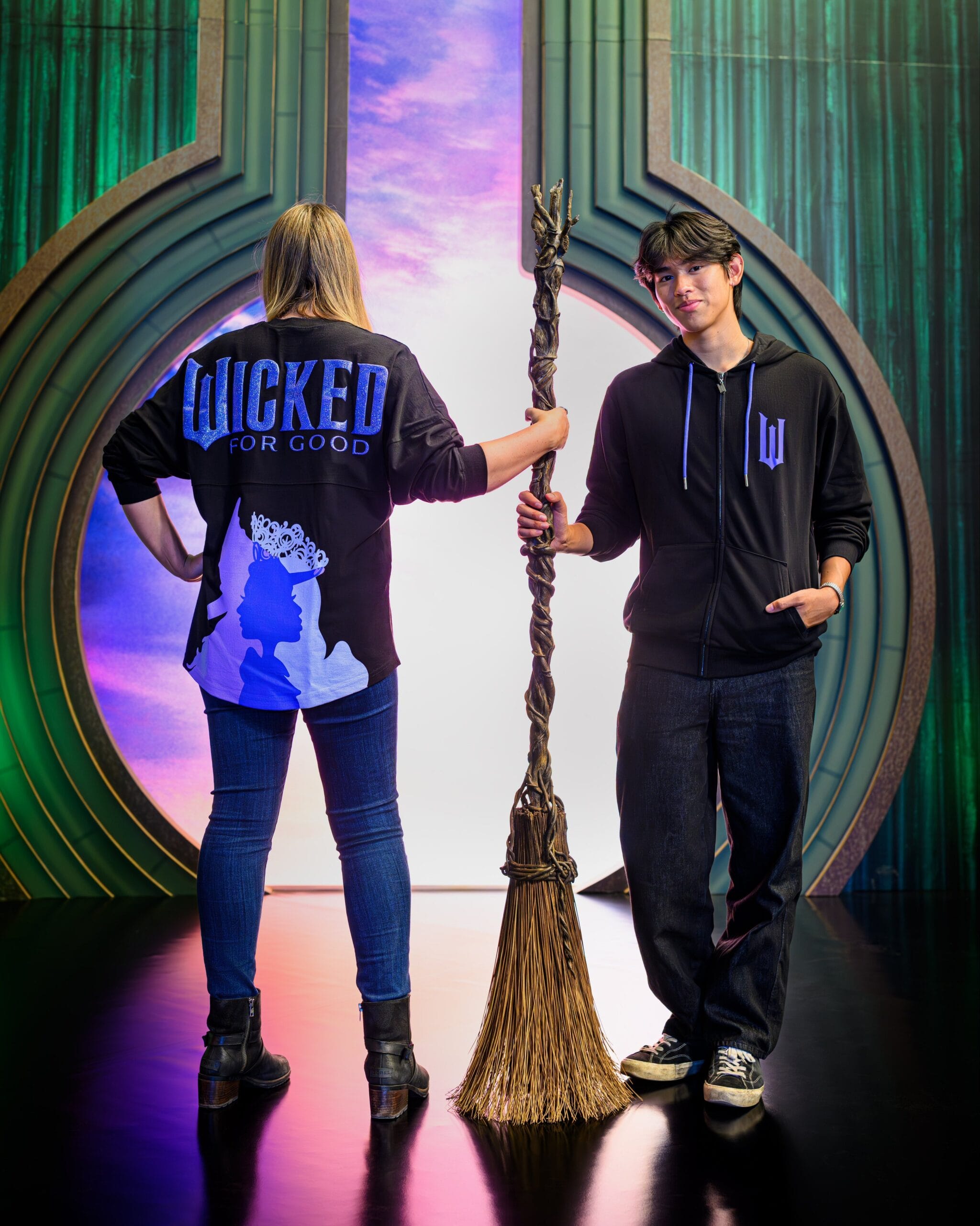 Two people pose at Universal Orlando—one in a "Wicked for Good" jacket, the other grins holding a broom, not a Disney castle.