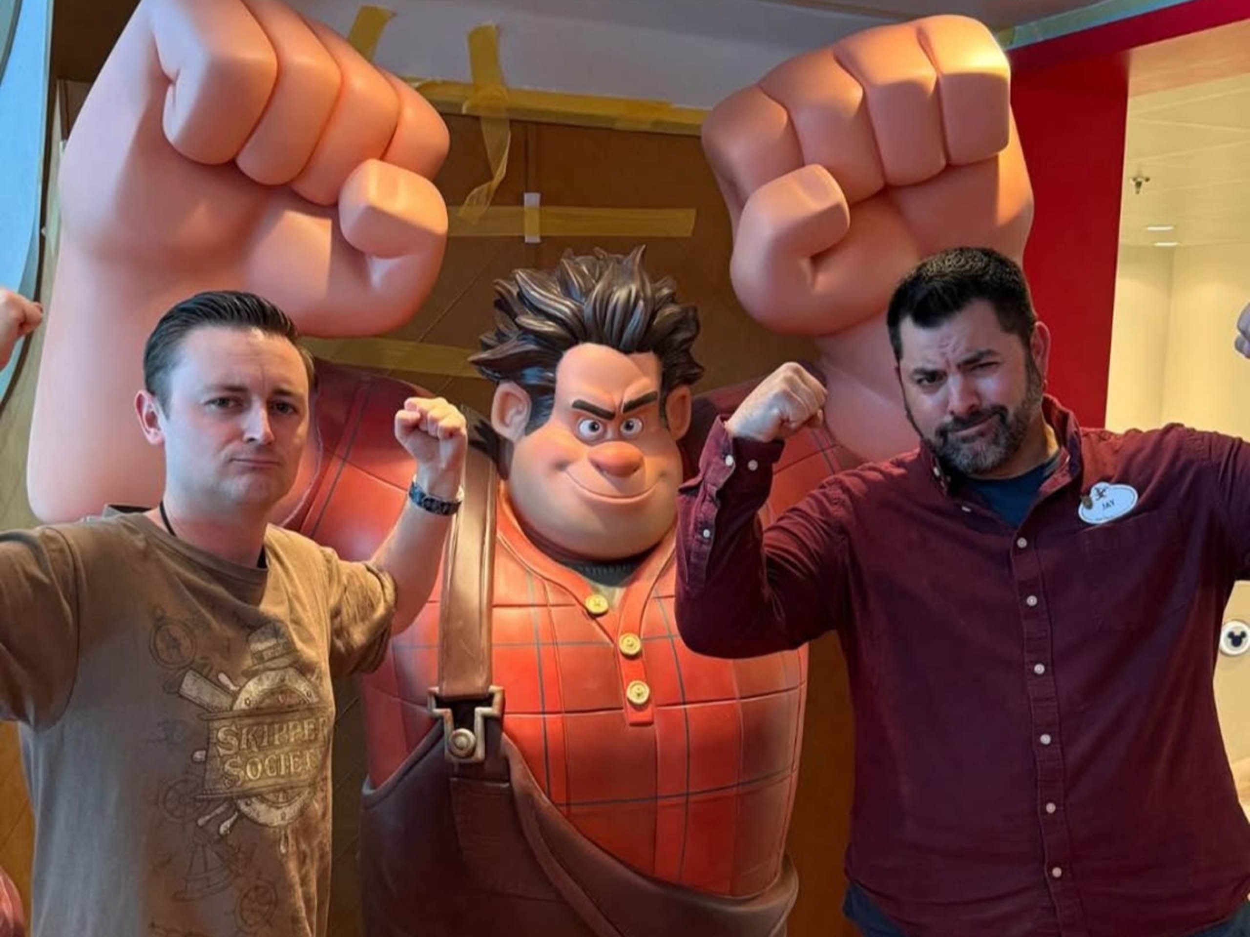Two men strike playful poses by a giant Wreck-It Ralph statue at a Disney park, embodying hero vibes with raised fists.