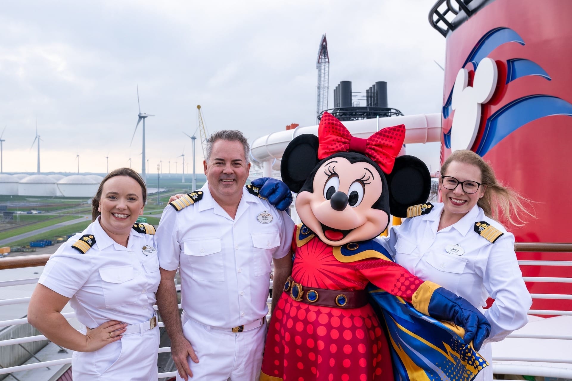 Three Disney Cruise staff in white uniforms pose with Minnie Mouse on deck; wind turbines spin behind—no castle in sight.