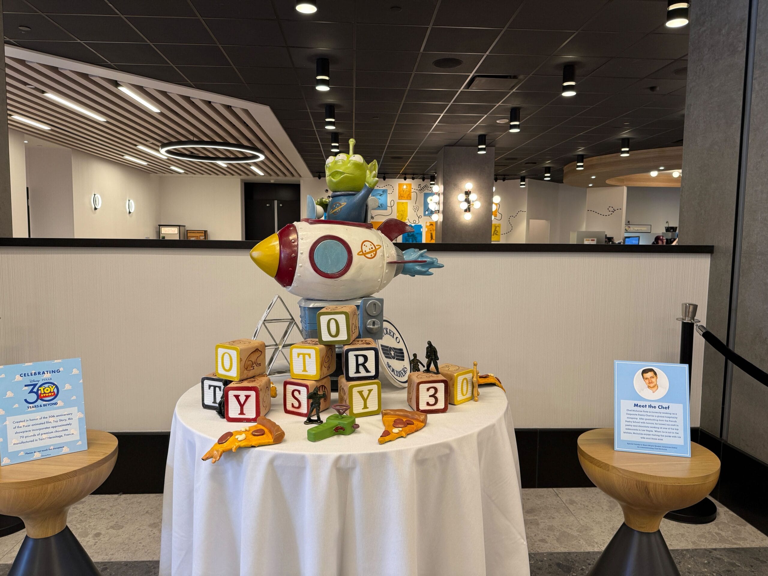 Toy Story-themed display with a rocket, alien figure, and letter blocks on a table in a Disney Park indoor area—no castle visible.