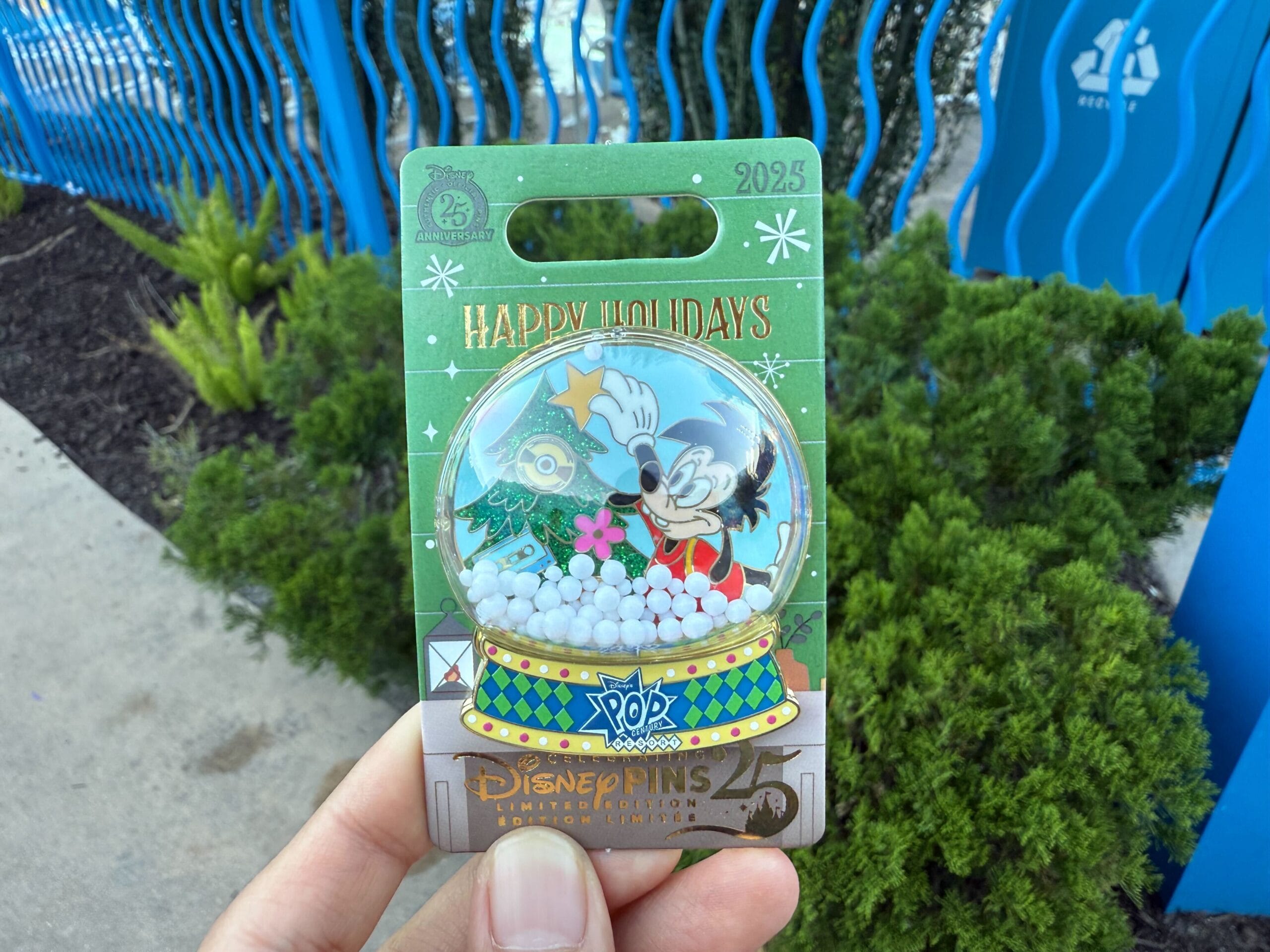 A hand displays a Walt Disney World holiday pin featuring Mickey in a snow globe, greenery, and a blue Resort Hotel fence behind—no castle visible.