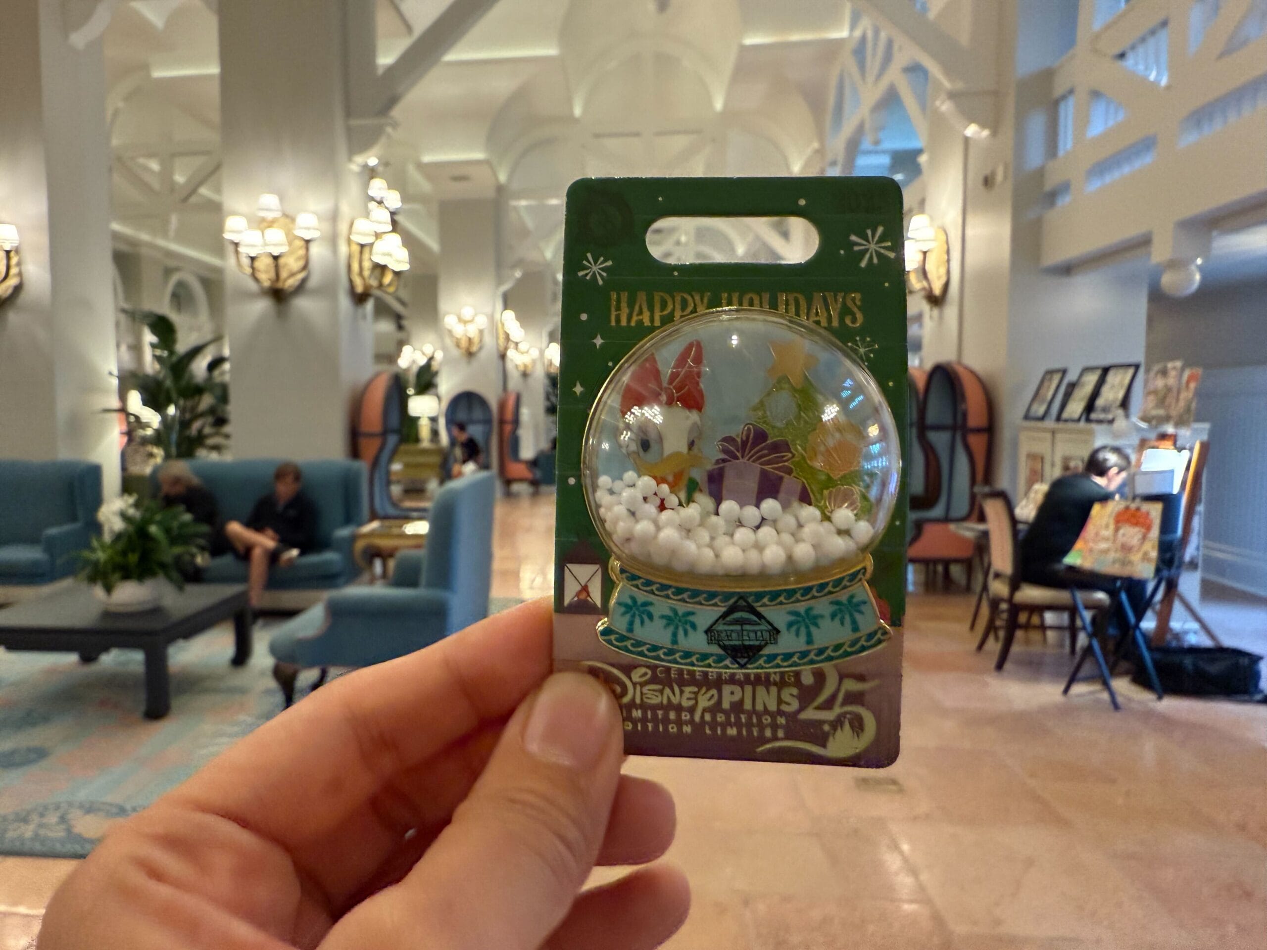 A hand holds a “Happy Holidays” Disney pin inside a sunny Walt Disney World resort lobby, guests lounging in the background.