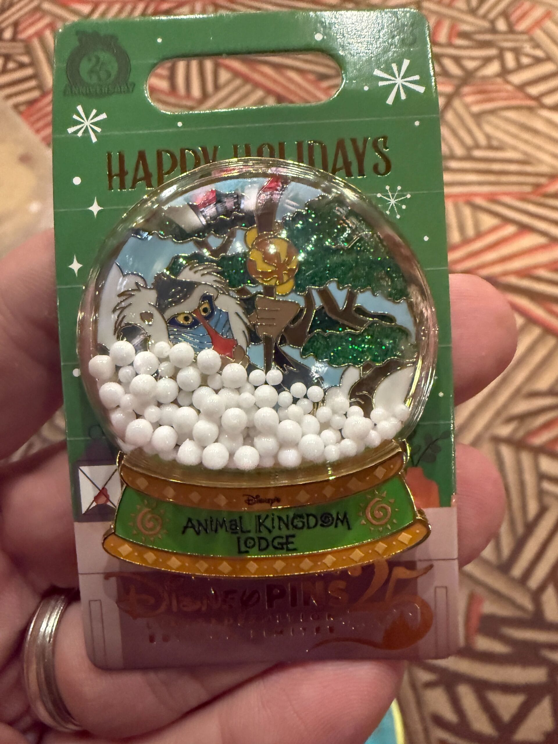A hand holds a Disney’s Animal Kingdom Lodge holiday snow globe pin with Rafiki; no castle like Cinderella’s or Sleeping Beauty’s shown.
