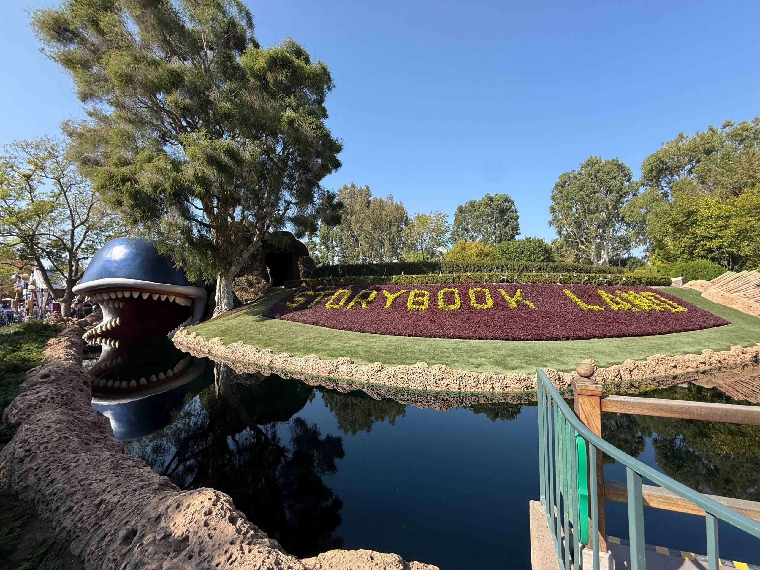 A flower bed spells "STORYBOOK LAND" by Monstro the whale’s mouth, next to a pond and trees, reminiscent of Disneyland’s classic ride.