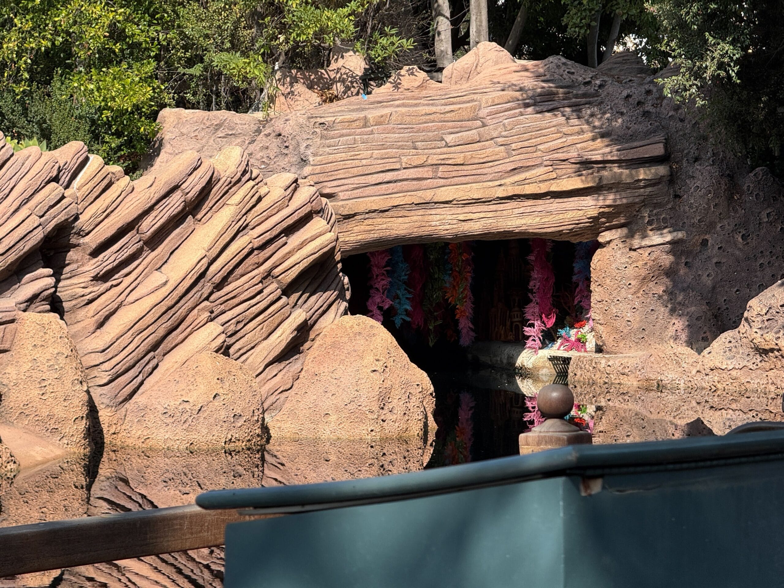 A rocky cave entrance, reminiscent of Disneyland's Storybook Land Canal Boats, adorned with vibrant flowers, reflected in tranquil water.