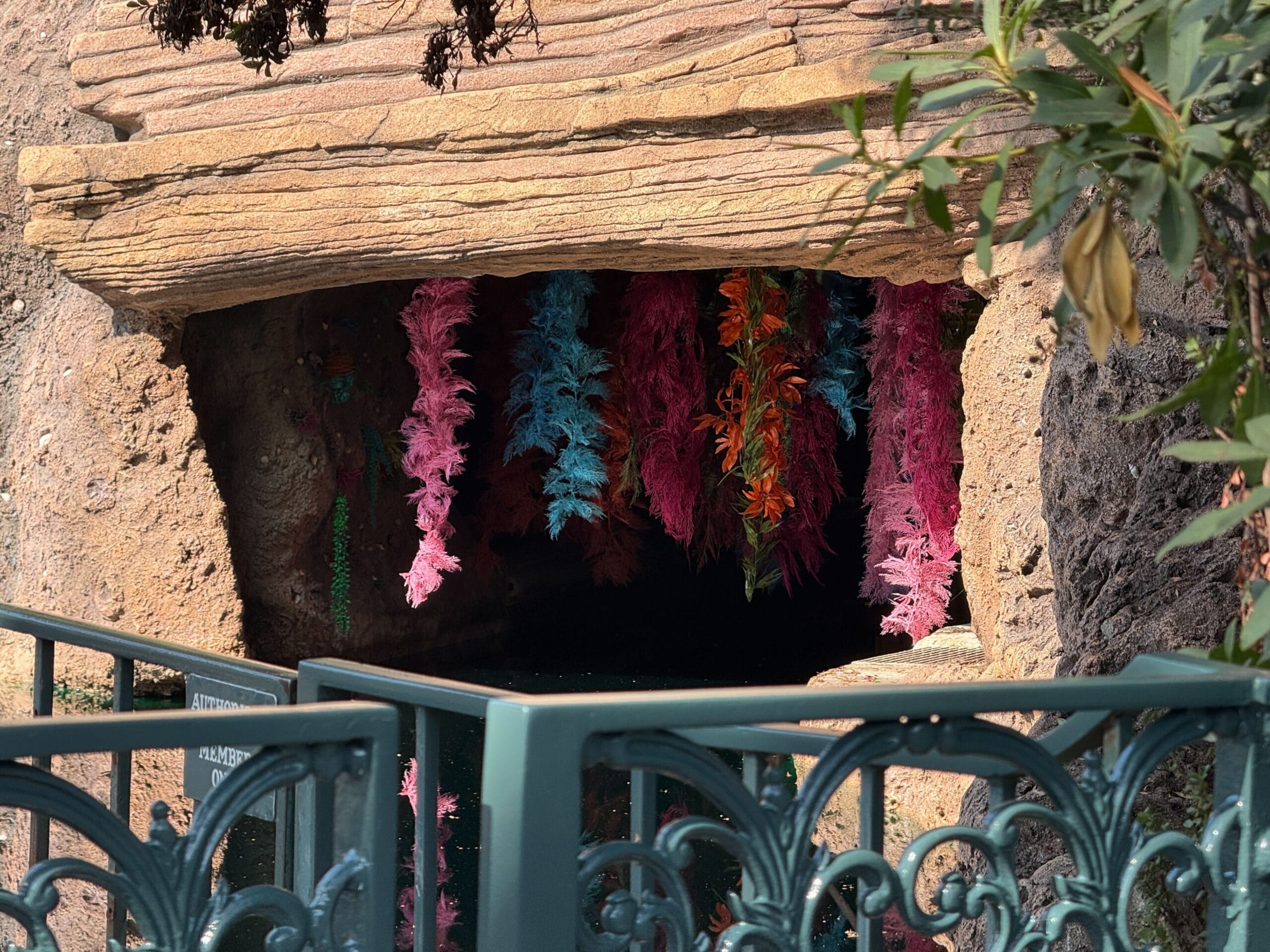A Disneyland cave entrance framed by vibrant artificial plants and a decorative green metal fence, no castle in this view.