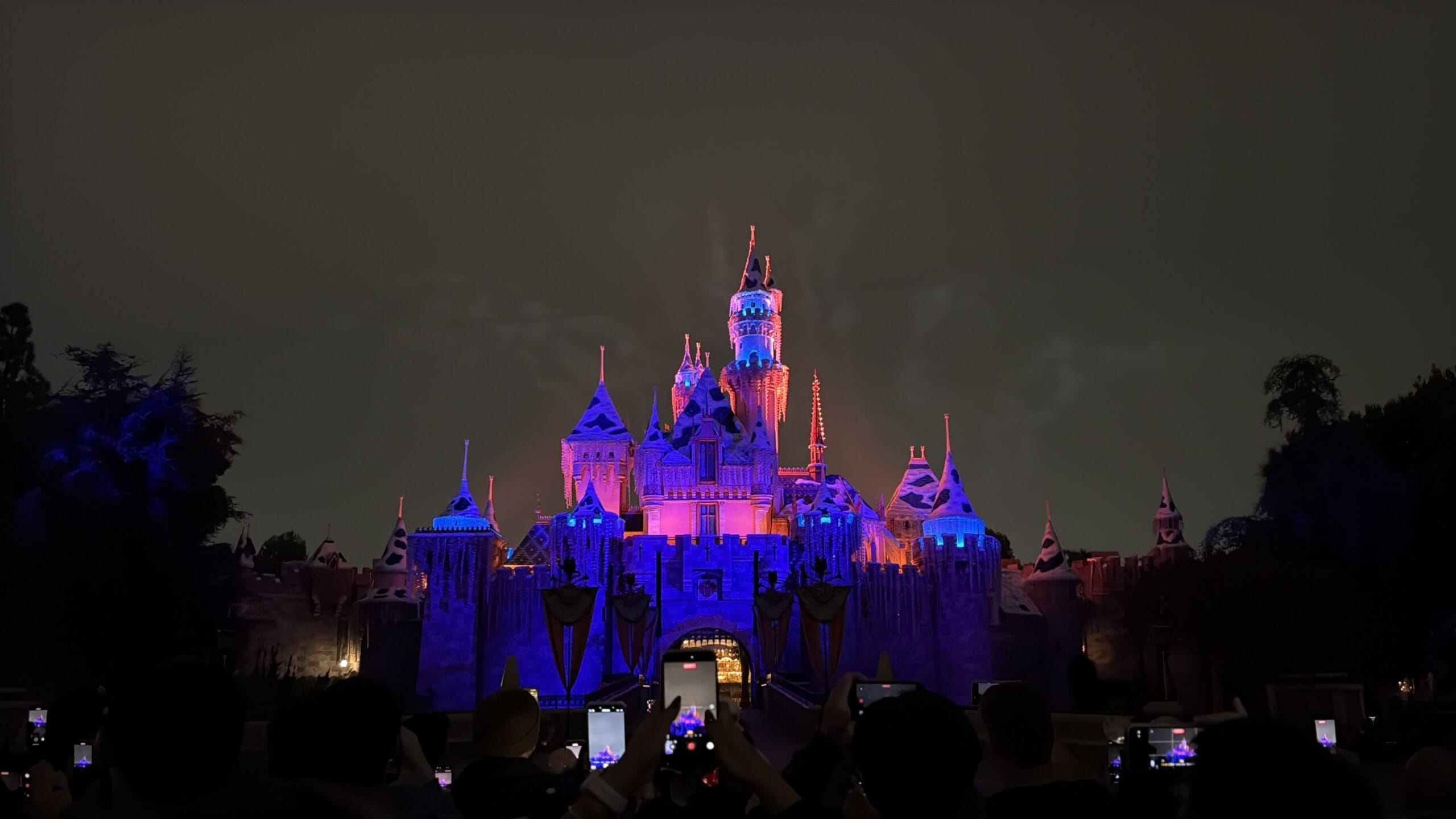 Sleeping Beauty Castle at Disneyland aglow in purple and blue for Wondrous Journeys, distinct from the taller Cinderella Castle in Florida.