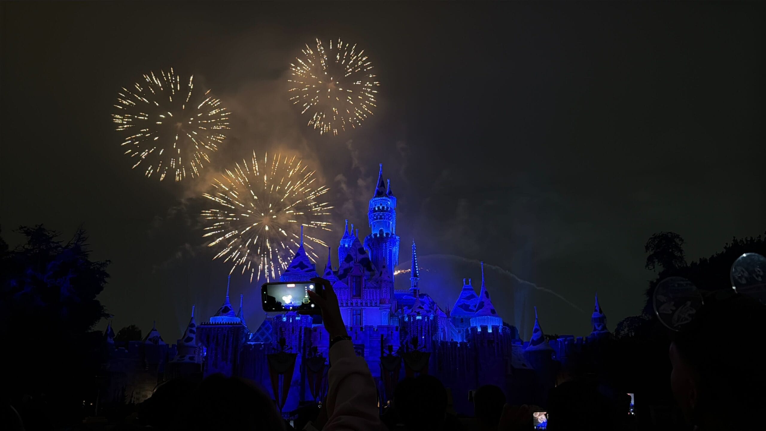 Fireworks sparkle above a blue-lit Disneyland castle, which resembles the original Anaheim design, as crowds film with their phones.