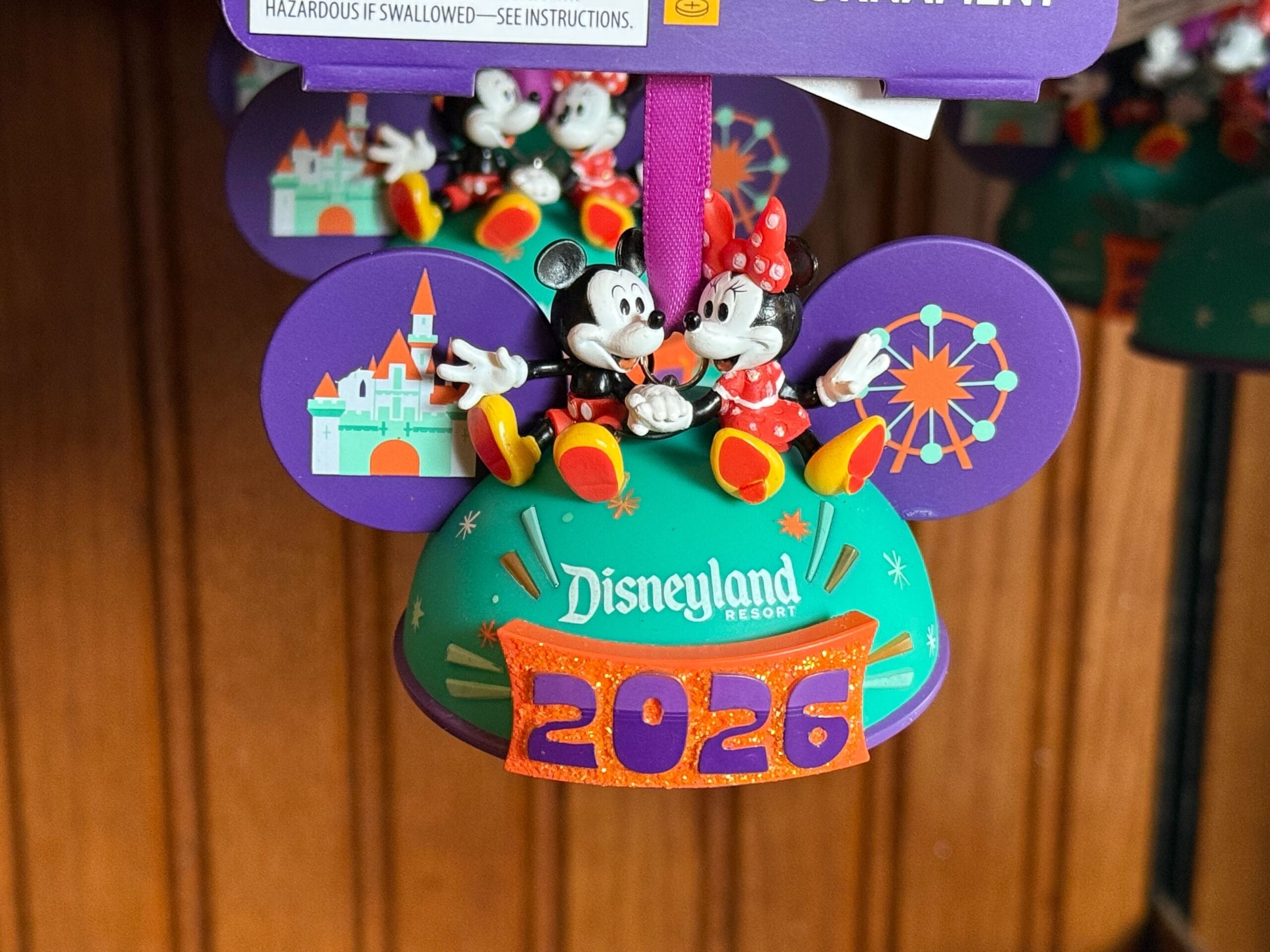 This ornament is adorable with Mickey and Minnie sitting on top, holding hands. The ears of the hat are purple with the Disneyland castle on the left and Pixar Pal-A-Round on the right. The bottom of the hat is cyan with the Disneyland Resort word mark above an orange banner that says 2026 in purple.