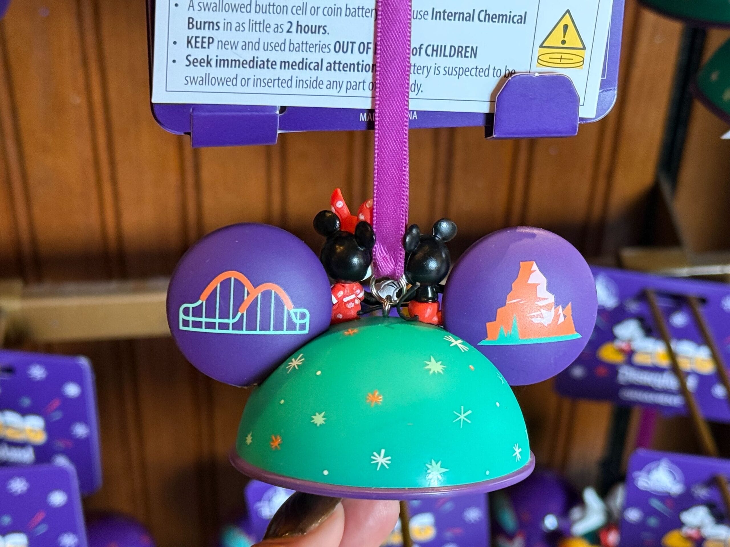 The back of the hat ornament is simple, with orange, white, and light cyan sparkles on the back. The left ear has the Incredicoaster on it, and the right has the Matterhorn. The ornament is hung by the magenta ribbon that is attached to the top.