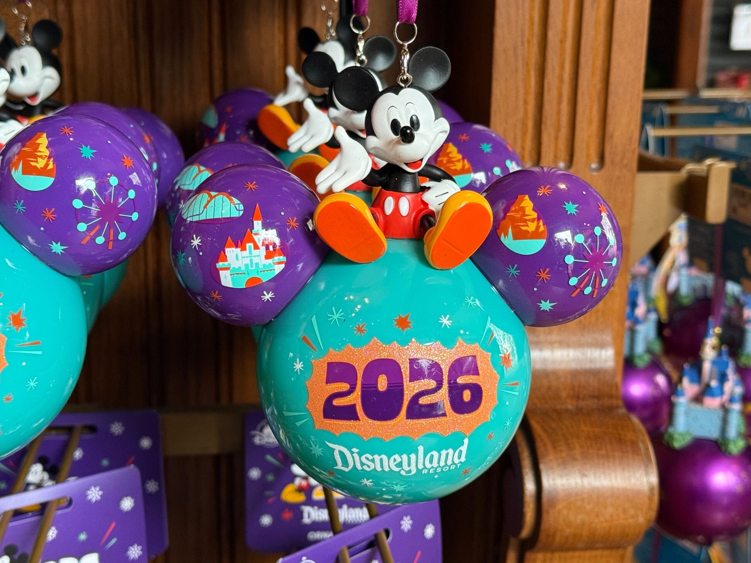 This ornament is cyan with the Disneyland Resort wordmark in white. Above is an orange bubble with 2026 written in purple. Mickey is sitting on top of this ornament with one hand on his hip and one hand held out. The left ear has the Incredicoaster and Disneyland castle, and the right has the Matterhorn and Pixar Pal-A-Round.