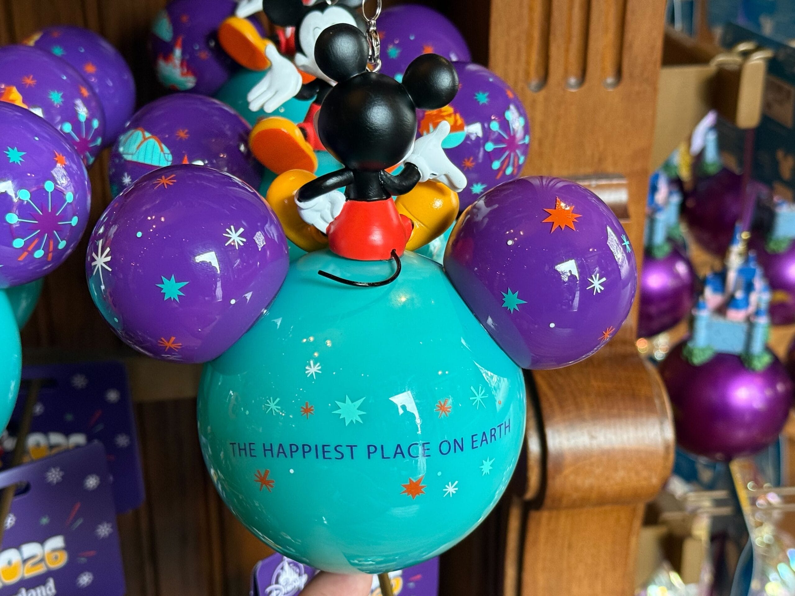 The back of the ornament on the cyan base says "The Happiest Place On Earth" written in purple. There are orange, cyan, and white sparkles all over the ornament.