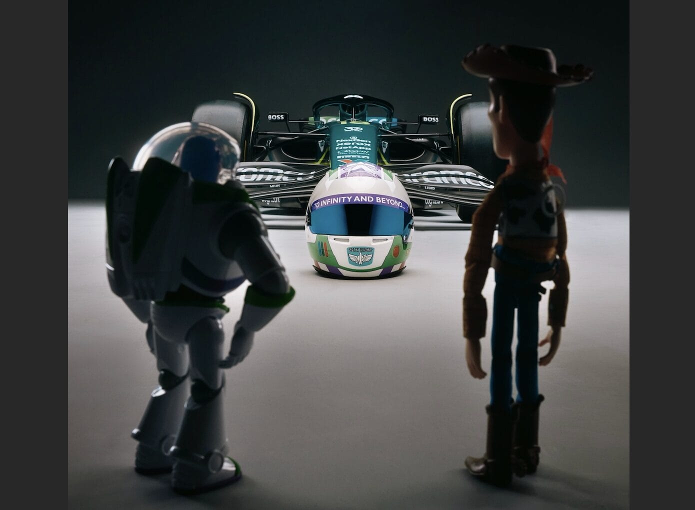 Buzz Lightyear and Woody stand before an Aston Martin F1 car reading "INFINITY AND BEYOND," much like a Disney park display.