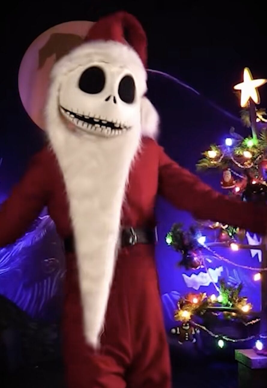 At a Disney park, Jack Skellington in an updated Santa suit poses by a vibrant Christmas tree during a festive meet and greet.