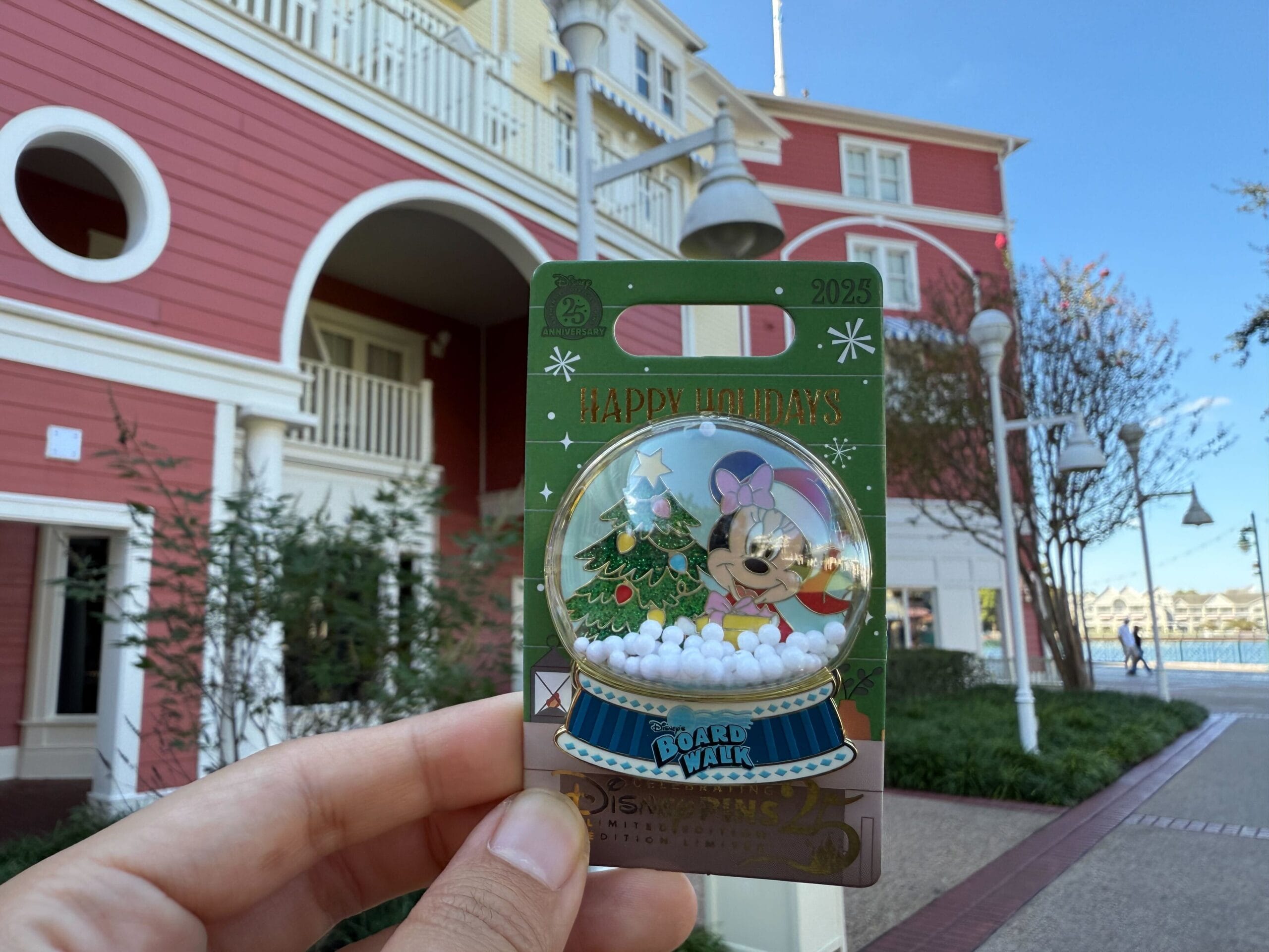 A hand holds a Walt Disney World holiday pin before a red building with round windows and white trim, not a Disney castle backdrop.