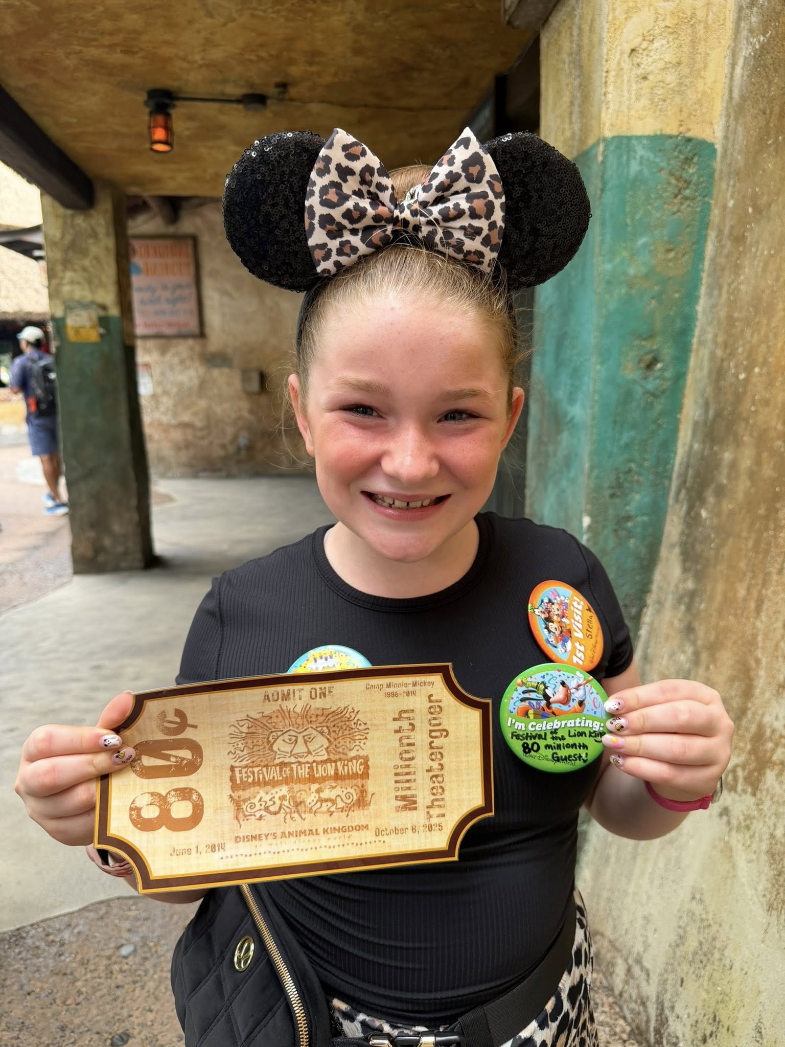 Smiling girl at Disney's Animal Kingdom wears Minnie ears, holding Lion King show ticket and celebration buttons, no castle in view.