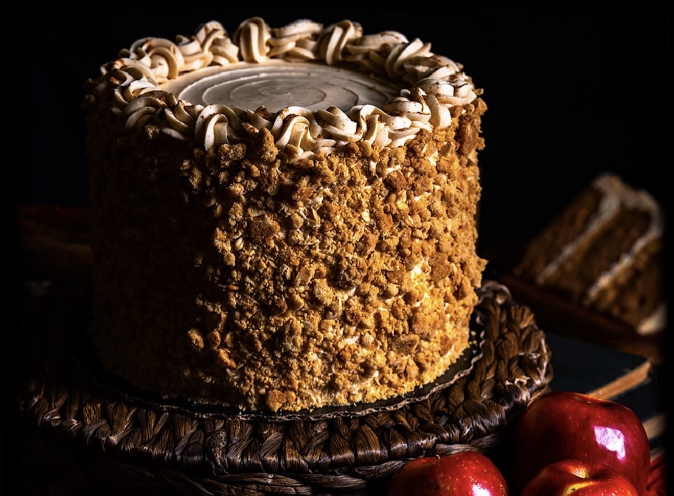 A tall, round crumb-coated cake from Gideon's Bakehouse sits on a woven tray beside shiny red apples, perfect for a Disney Parks treat.