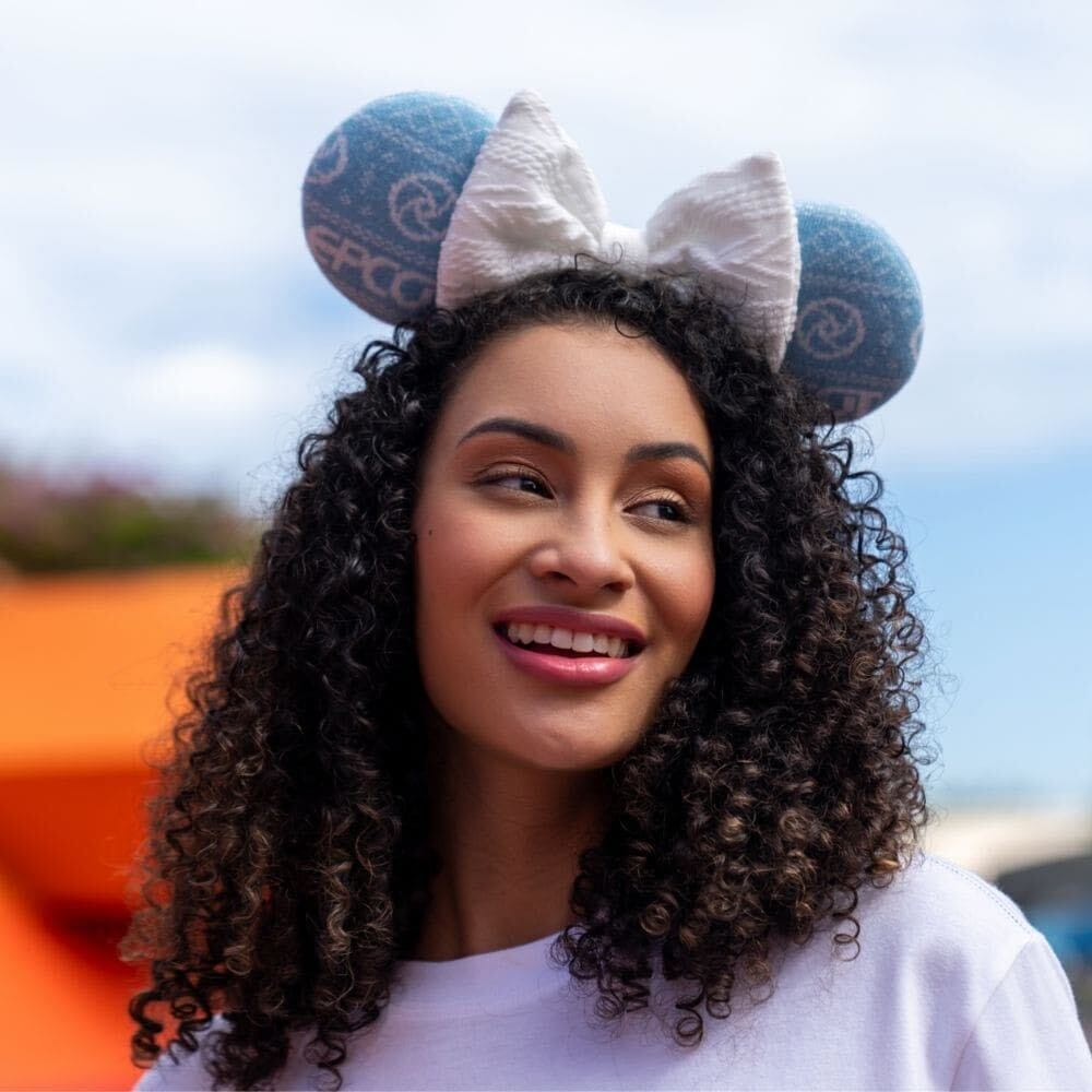 Smiling woman with curly hair wears blue Epcot mouse ears from the 2025 collection, posing outdoors at a Disney park.