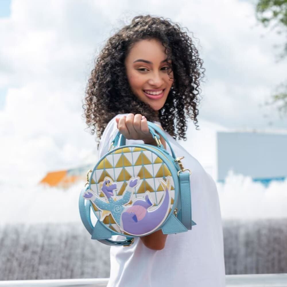 Smiling woman by an EPCOT fountain holds a round purse featuring Figment, the purple dragon, highlighting unique park merchandise.