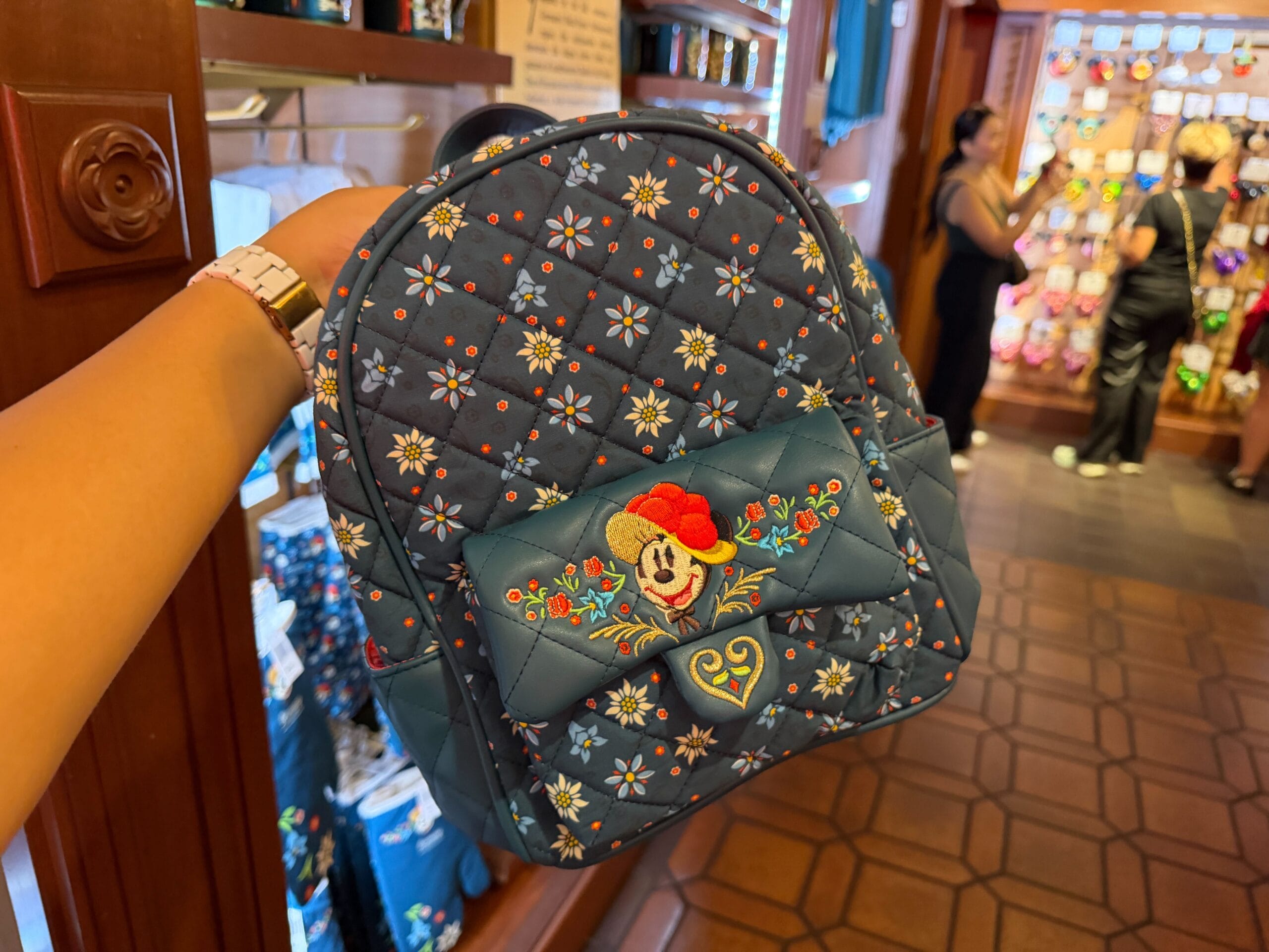 A hand holds a quilted blue Minnie Mouse backpack with floral designs inside a Disney park store in Germany, no castle visible.