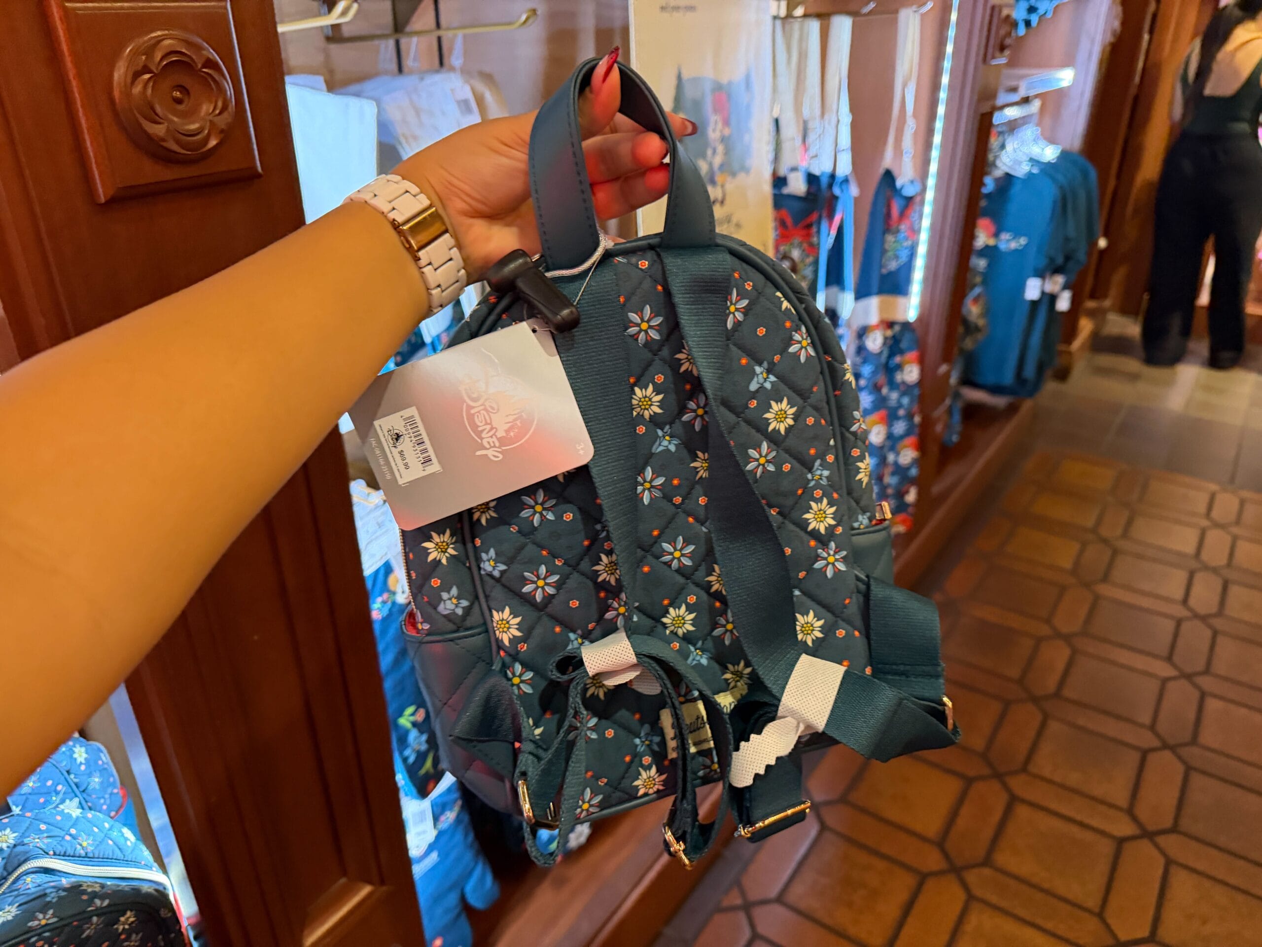 Guest holding a blue floral backpack with tags in a Disney park shop, surrounded by merch themed after Germany’s Black Forest.