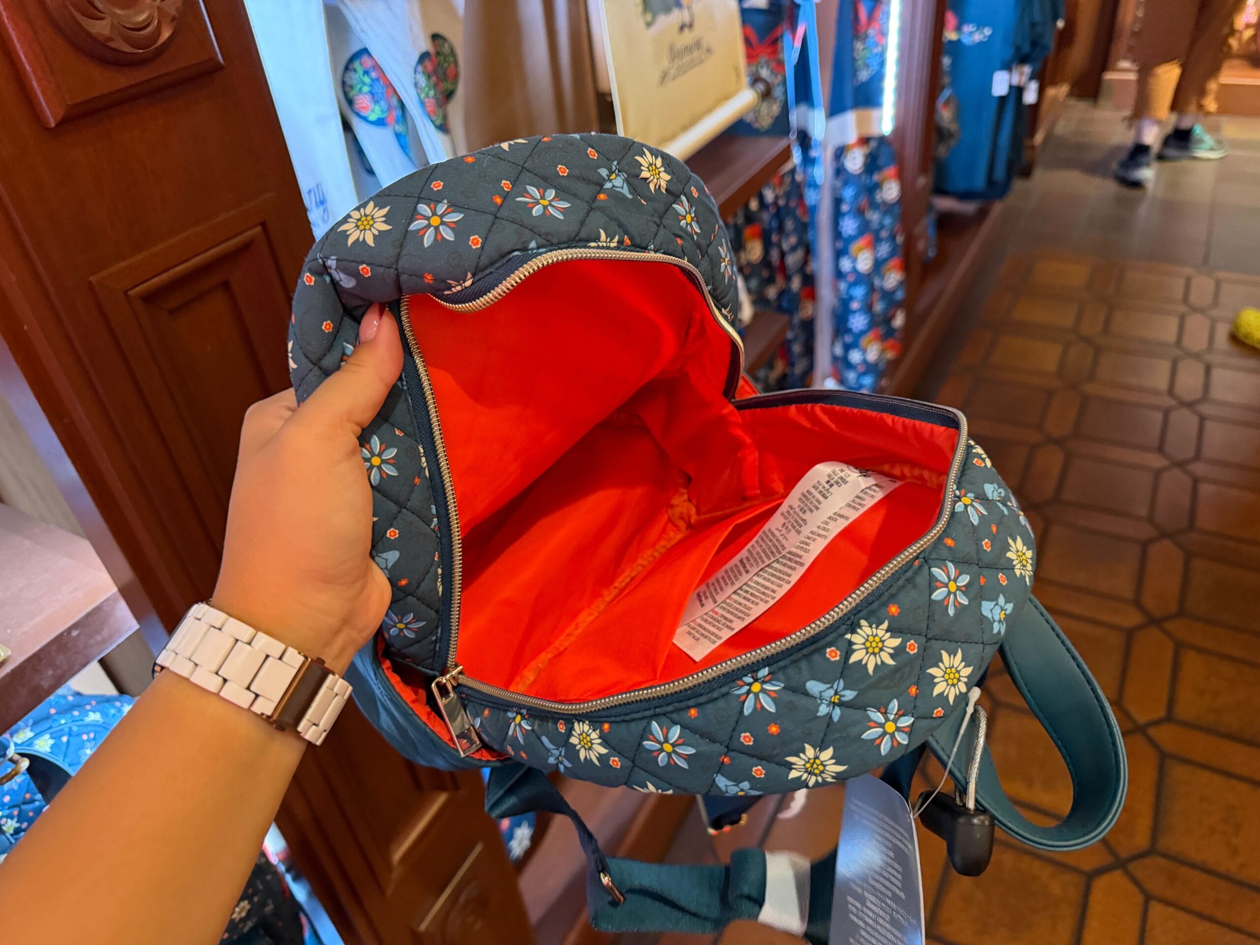 A hand opens a floral backpack with a vivid orange lining—Disney Parks merch inspired by Germany’s Black Forest, not castle-themed.