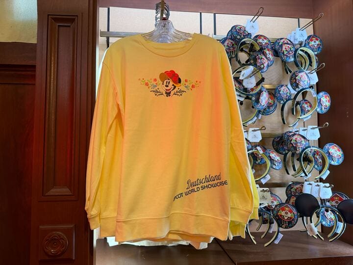 Yellow sweatshirt features floral Mickey, "Deutschland Epcot World Showcase" text, Disney Park merch near classic Mickey ear headbands.