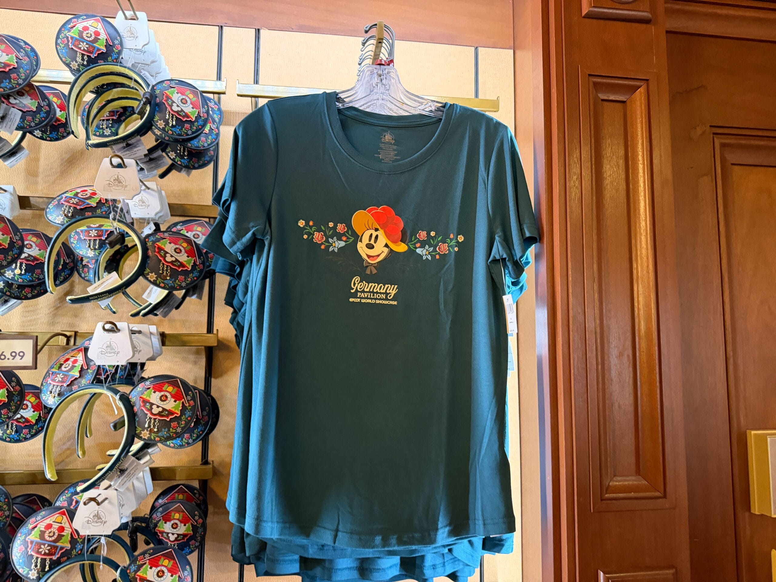 Teal tee featuring Mickey Mouse in a red hat, flowers, and "Germany" text—Black Forest Disney parks merch near headbands display.
