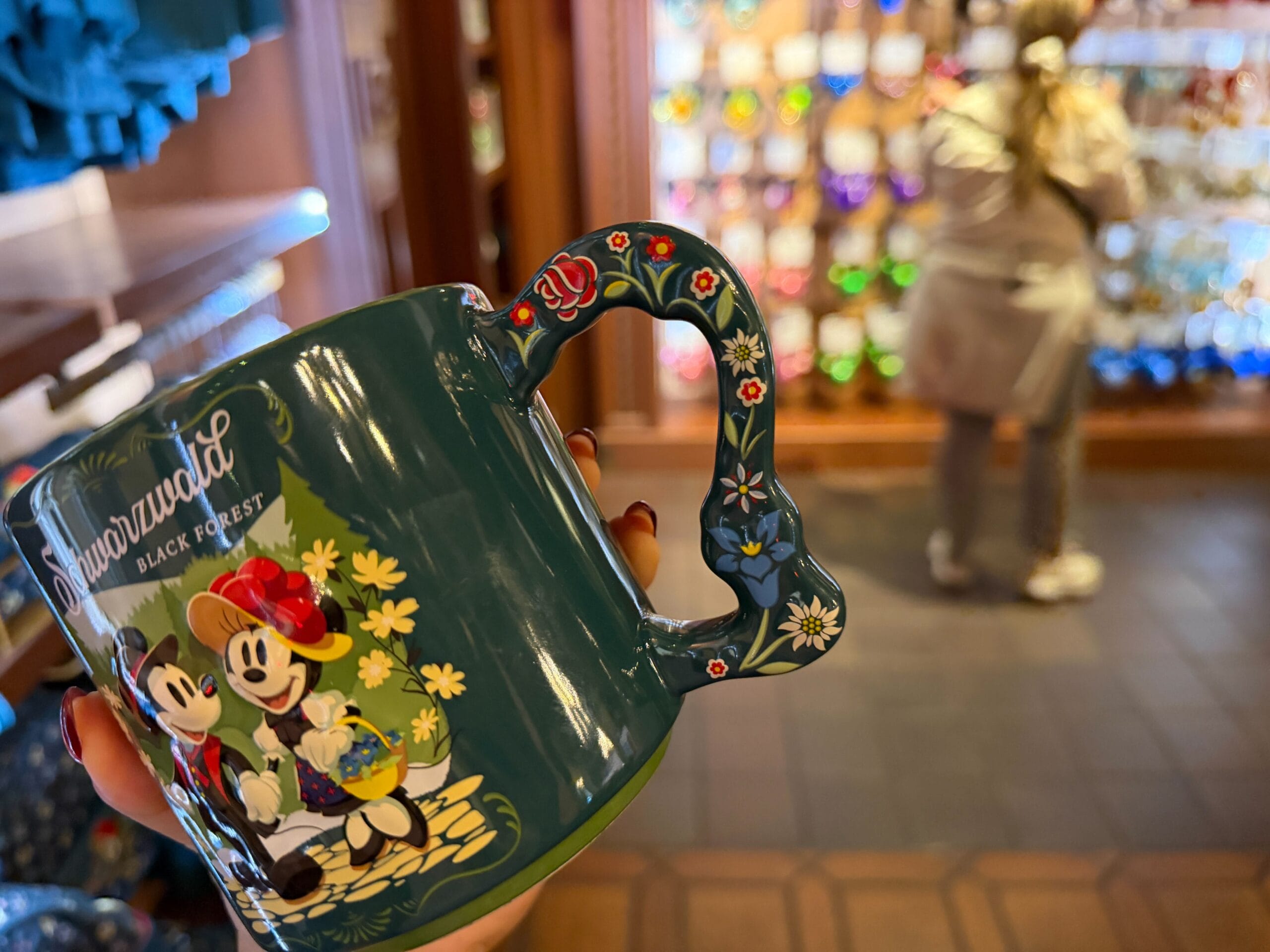 A hand holds a Germany-themed Minnie and Mickey Mouse mug inside a Disney park shop, with blurred shelves and a guest in back.