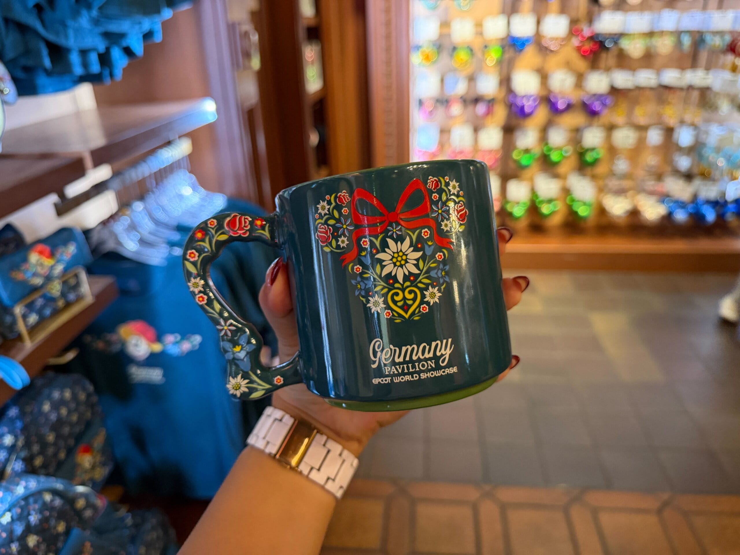 A hand holds a floral Germany Pavilion mug in a Disney shop, surrounded by vibrant Black Forest-inspired merchandise displays.