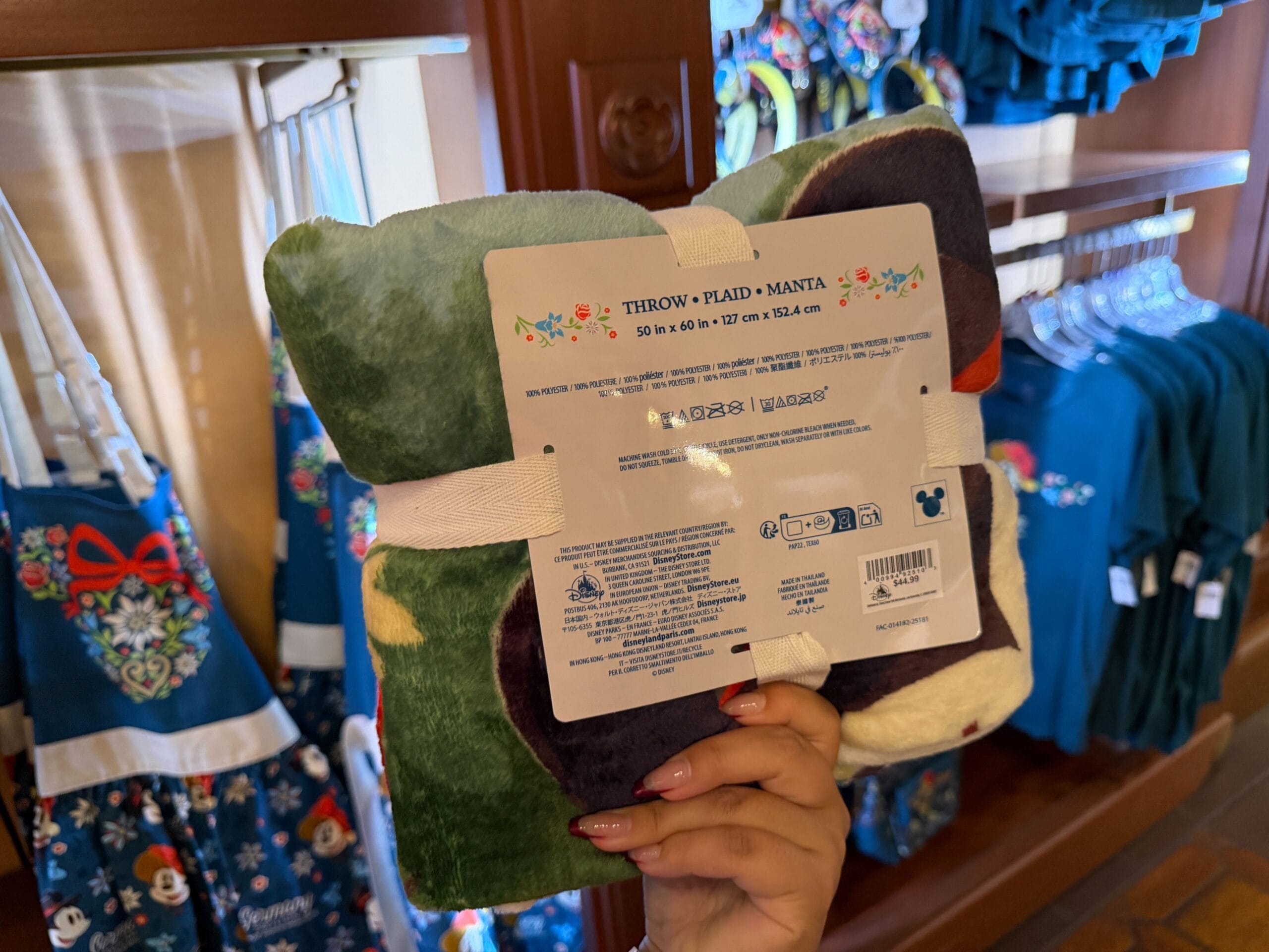 A hand holds a folded Disney blanket in a park shop, with themed aprons and shirts visible behind—classic Disney Parks merch.