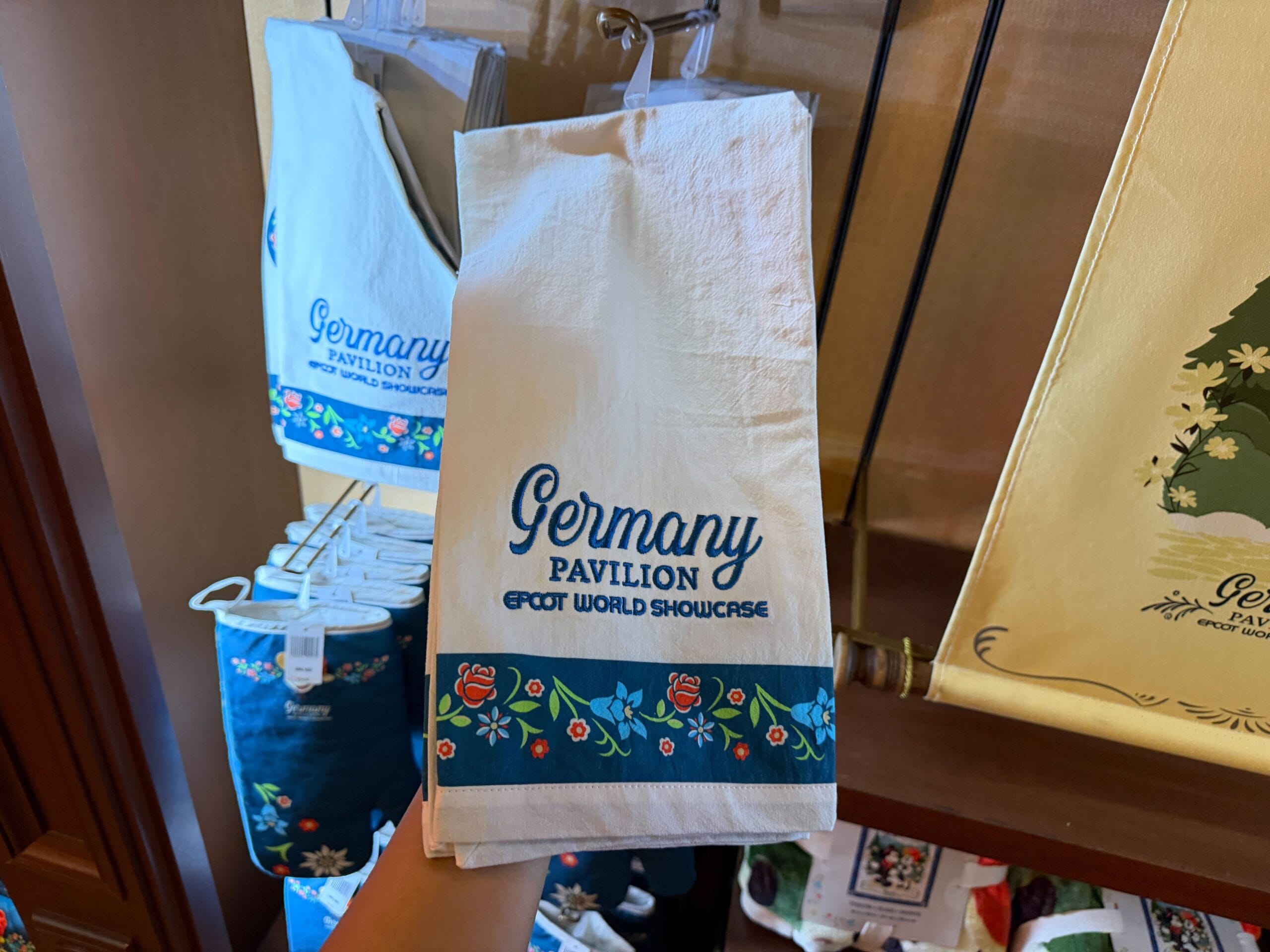A hand holds Germany Pavilion Epcot merch—a towel with castle-inspired Black Forest floral design, nodding to Disney’s global parks.
