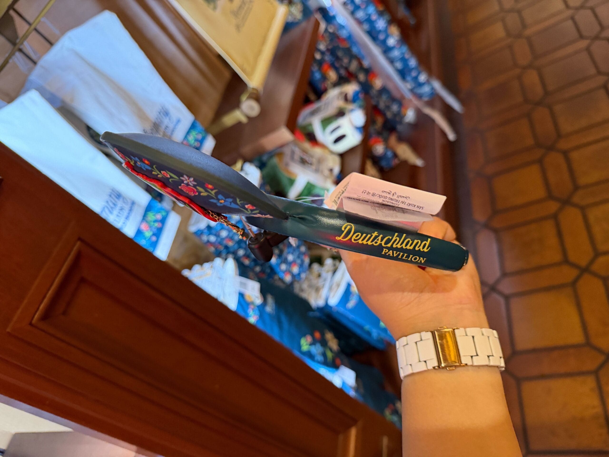 A guest at EPCOT’s Germany Pavilion holds a “Deutschland Pavilion” fan above Bavarian-themed Disney merchandise on display.