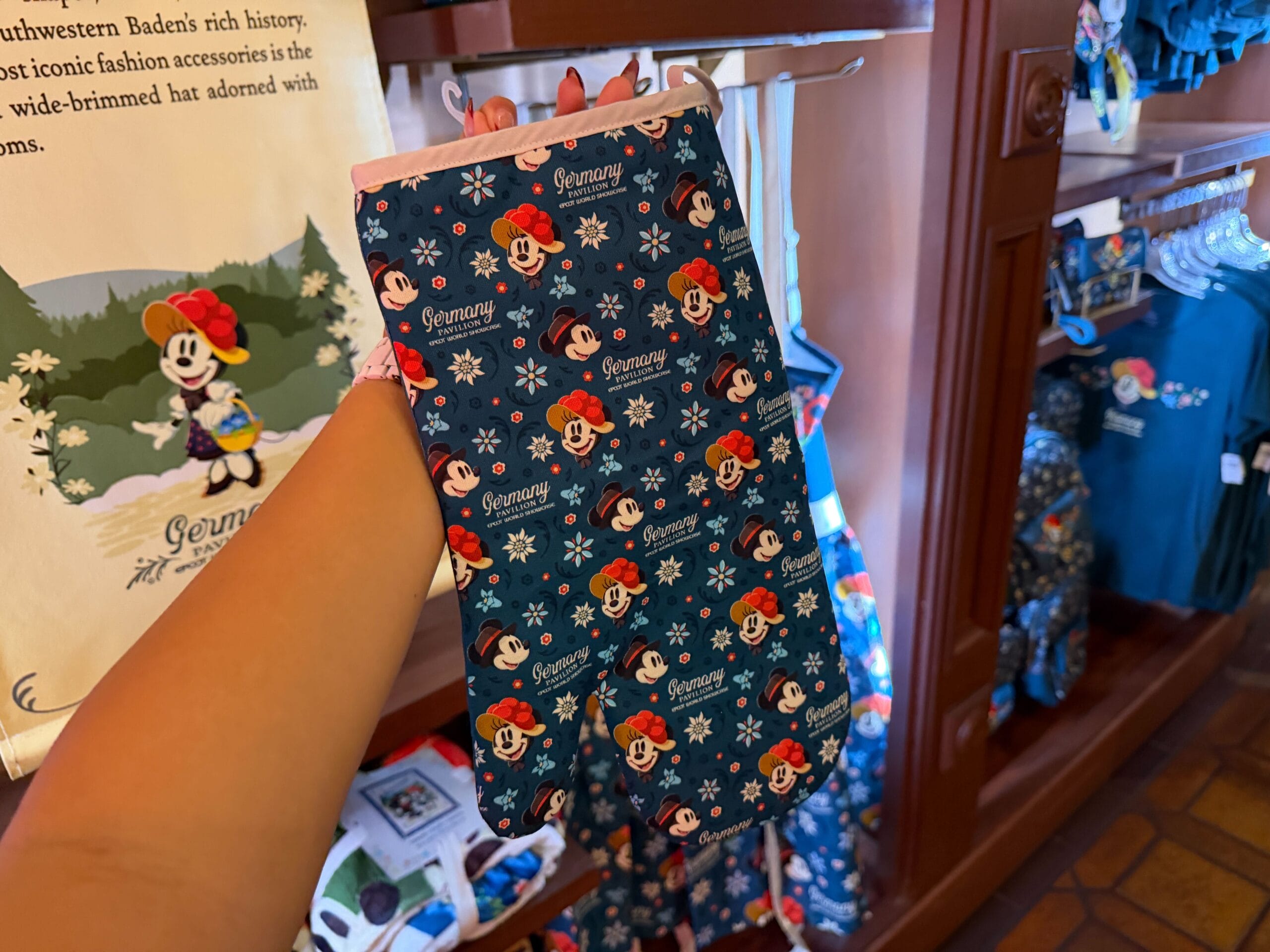 A hand holds a blue oven mitt with Minnie Mouse, snowflakes, and "Germany," spotted in a Disney park souvenir shop.