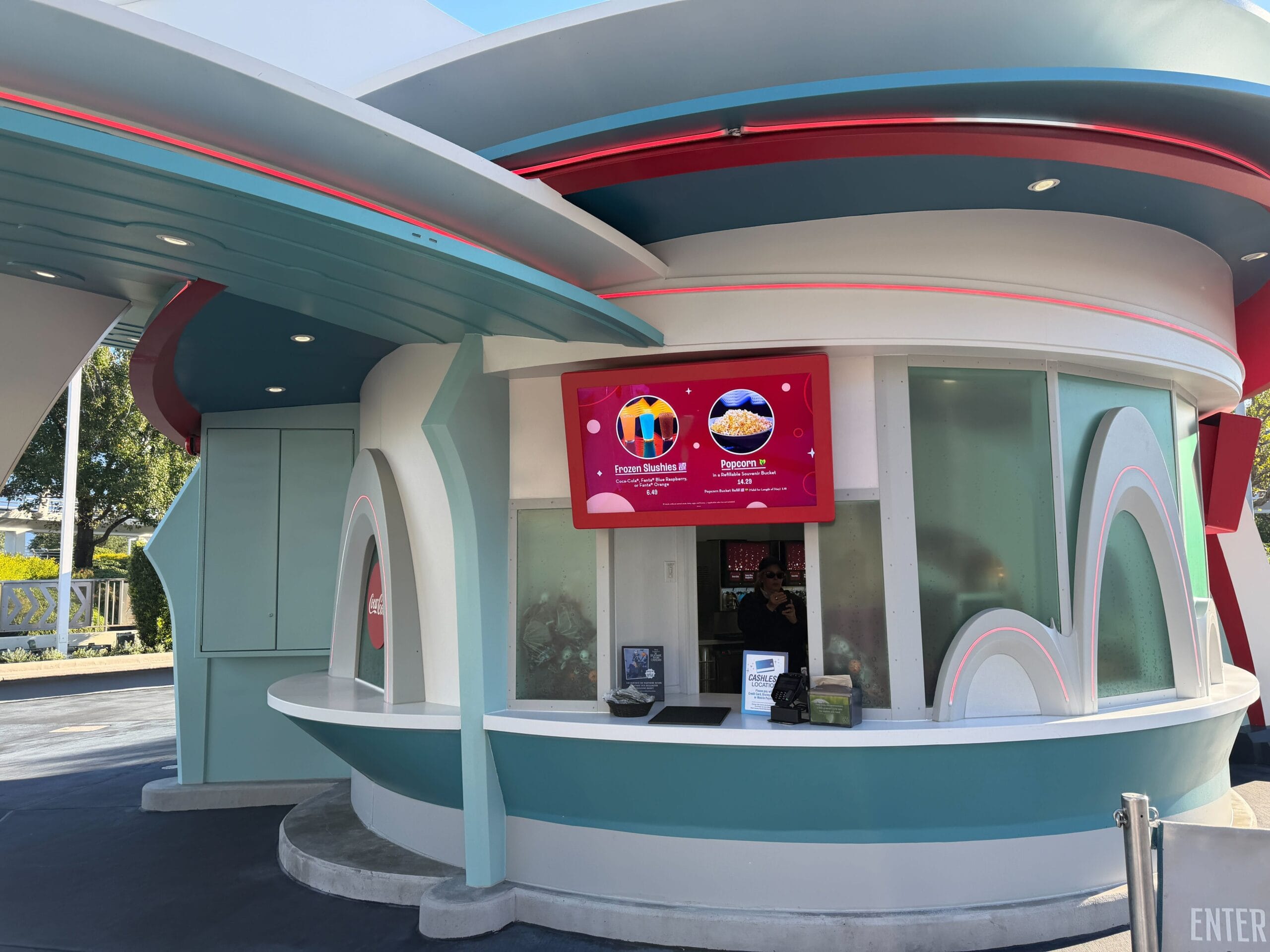 A retro-futuristic food kiosk in a Disney park serves Astro Fizz snacks with a digital menu, outdoors on a sunny day.