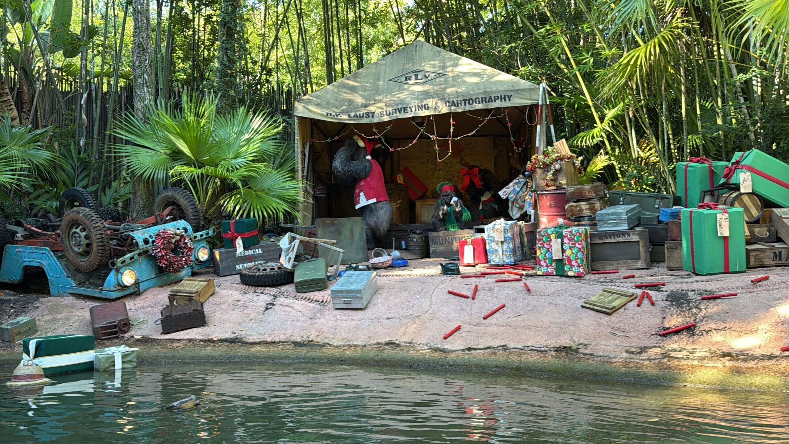A festive Jingle Cruise scene at a Disney park—tent, gorilla, gifts, and an overturned jeep beside the river jungle.