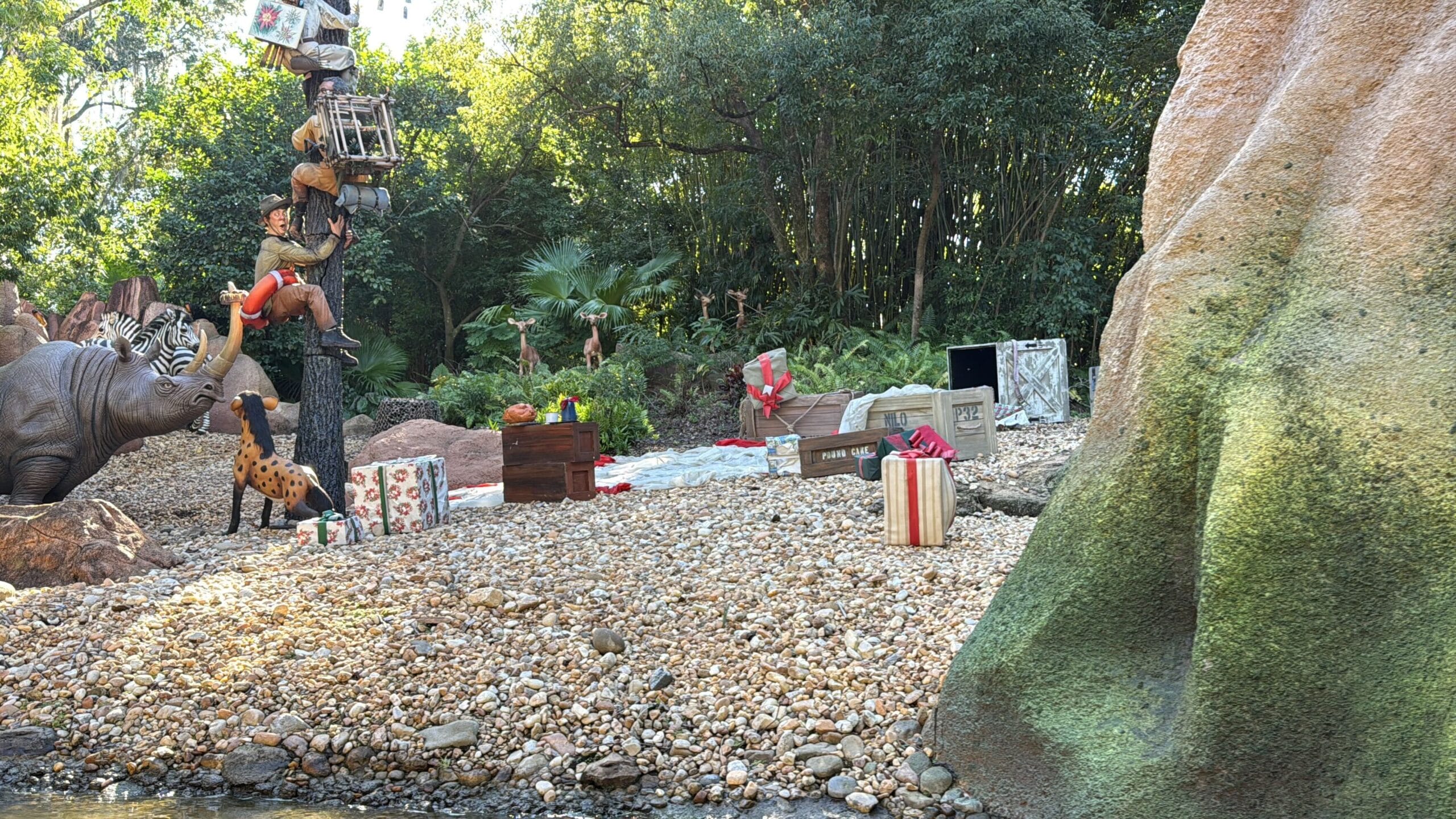 Guests and animals gather on a rocky riverbank with Jingle Cruise holiday décor