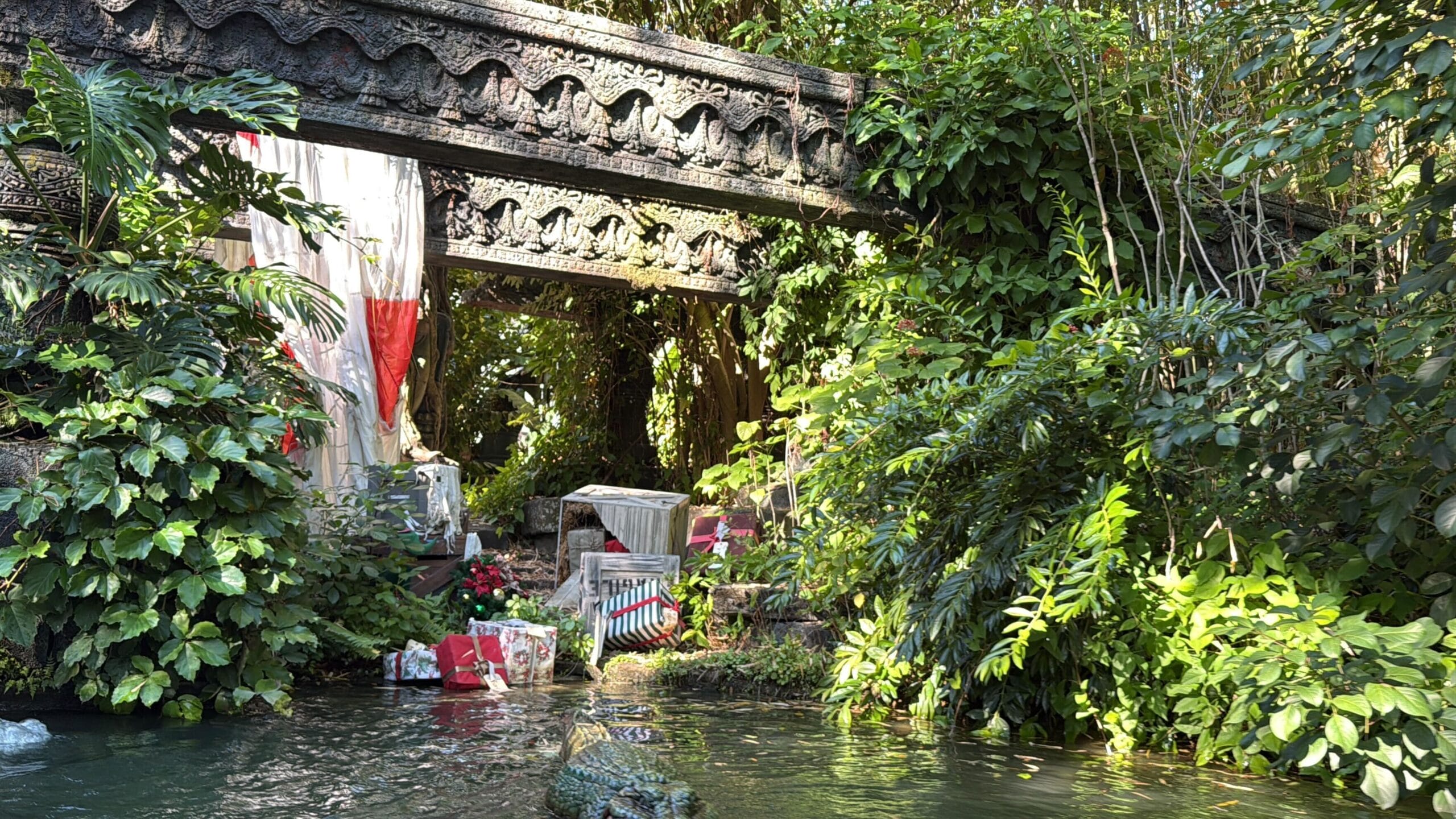 A decorative stone bridge crosses lush jungle foliage at Magic Kingdom’s Jingle Cruise, holiday gifts visible along the shore.