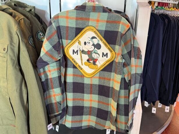 A plaid shirt from the Mickey Mouse Collection with a back patch, displayed on a rack in a Disney park merchandise store.