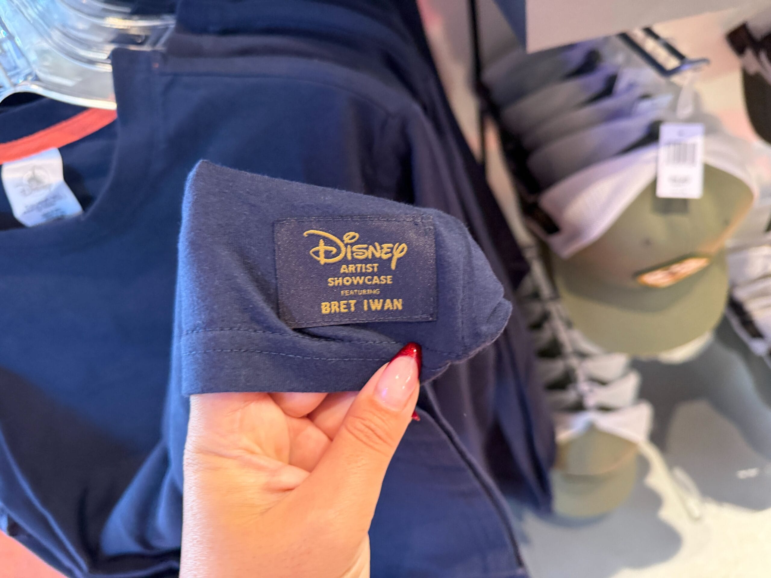 A hand holding a blue shirt from the Mickey Mouse Collection Expedition by Bret Iwan at a Disney Park, no castle in view.