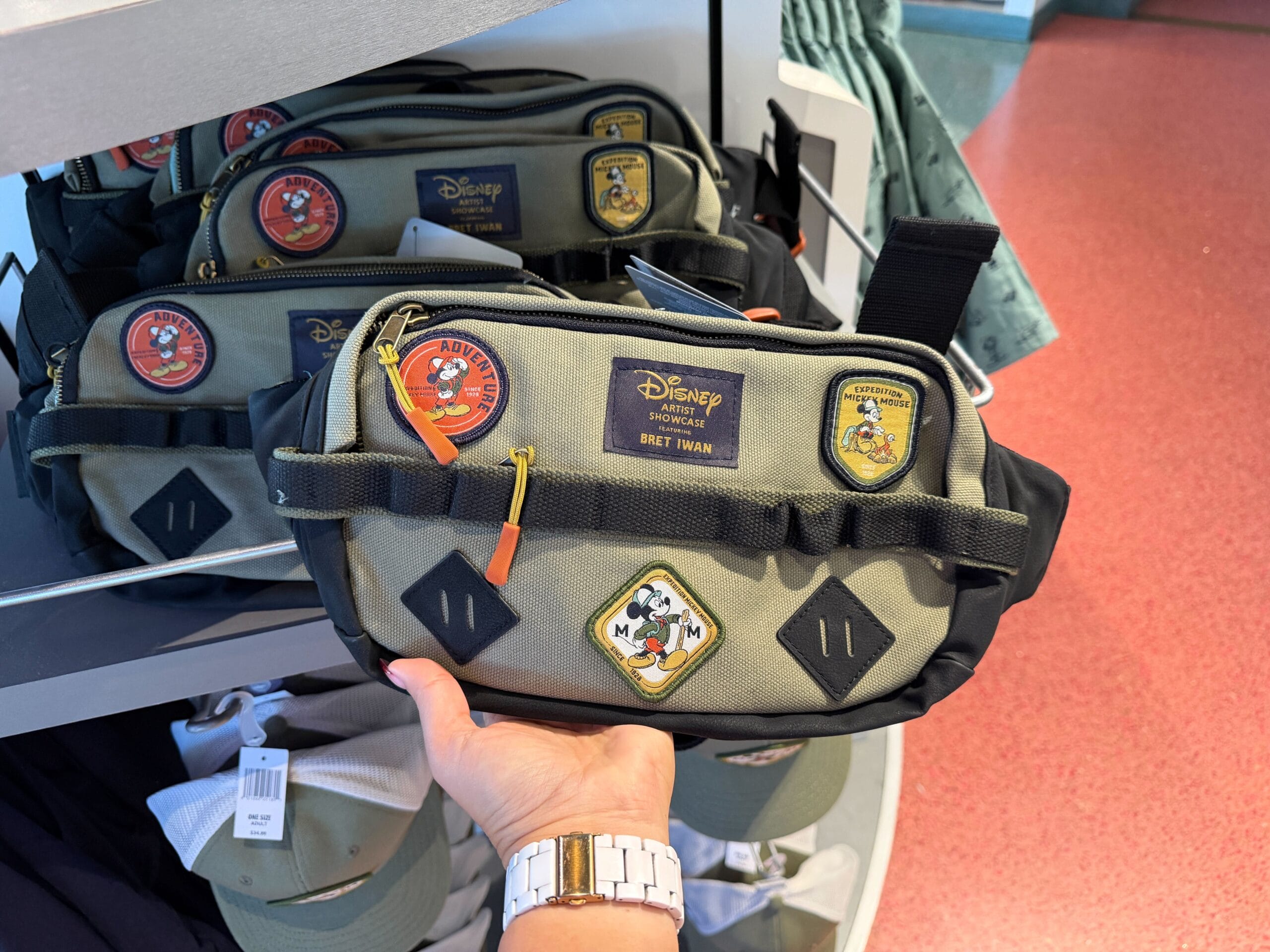 A hand displays an Expedition Mickey Mouse belt bag with adventure-themed patches, set on a Disney Park store shelf.
