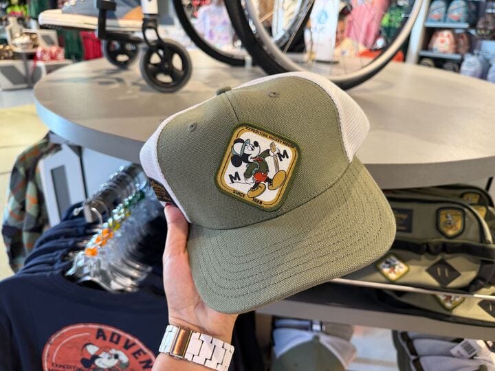 A guest holds a green and white Mickey Mouse Collection cap inside a Disney Park shop, with themed souvenirs visible behind.